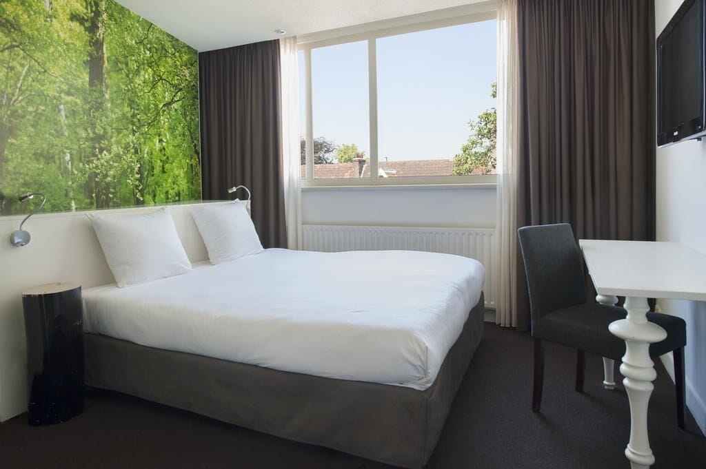 A hotel room with a bed, a desk, a chair, a window with curtains, and a wall with a nature-themed mural.