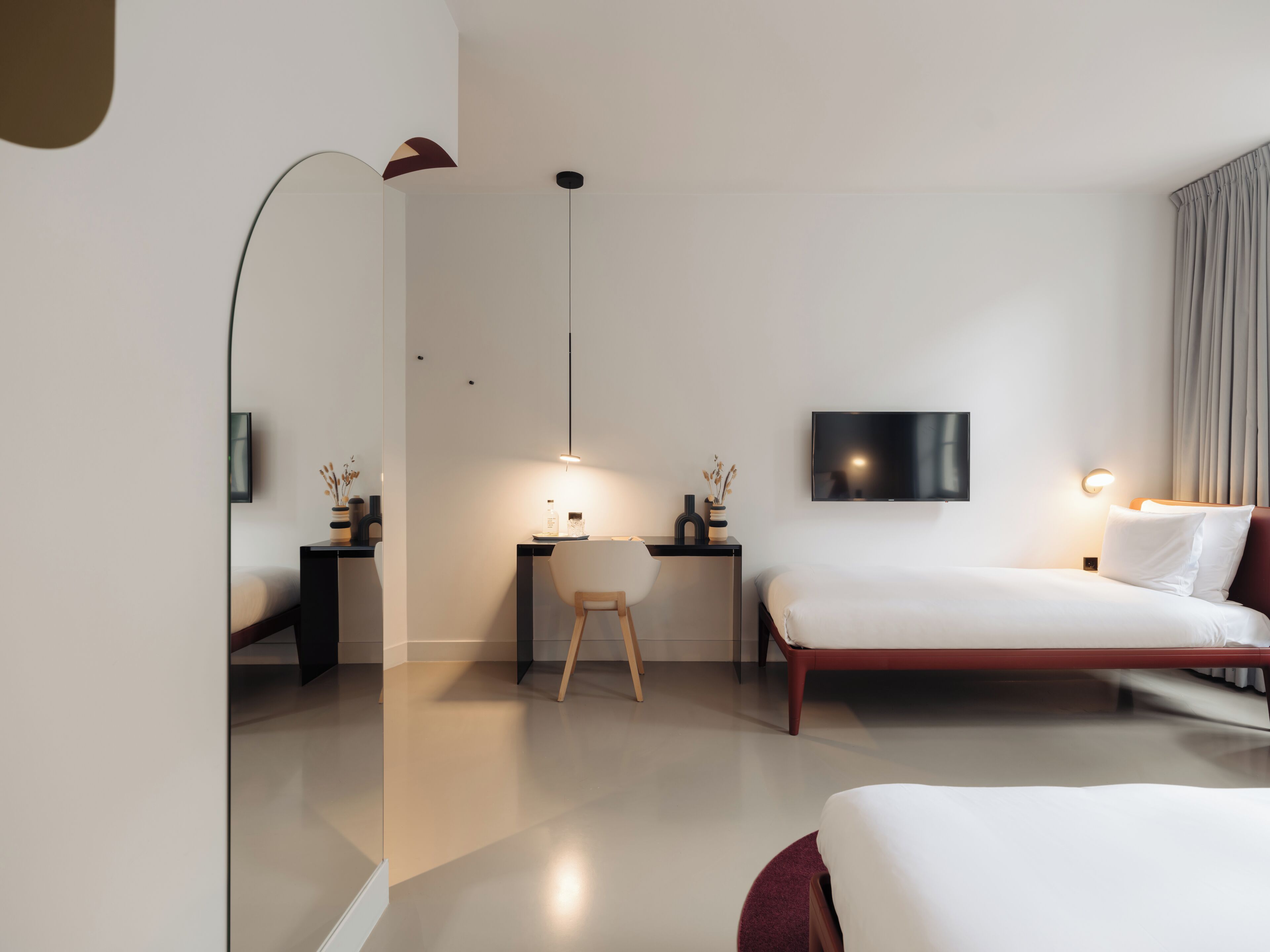 Quarto de hotel moderno com uma cama grande, uma escrivaninha com cadeira, uma TV fixada na parede e uma luminária pendente.