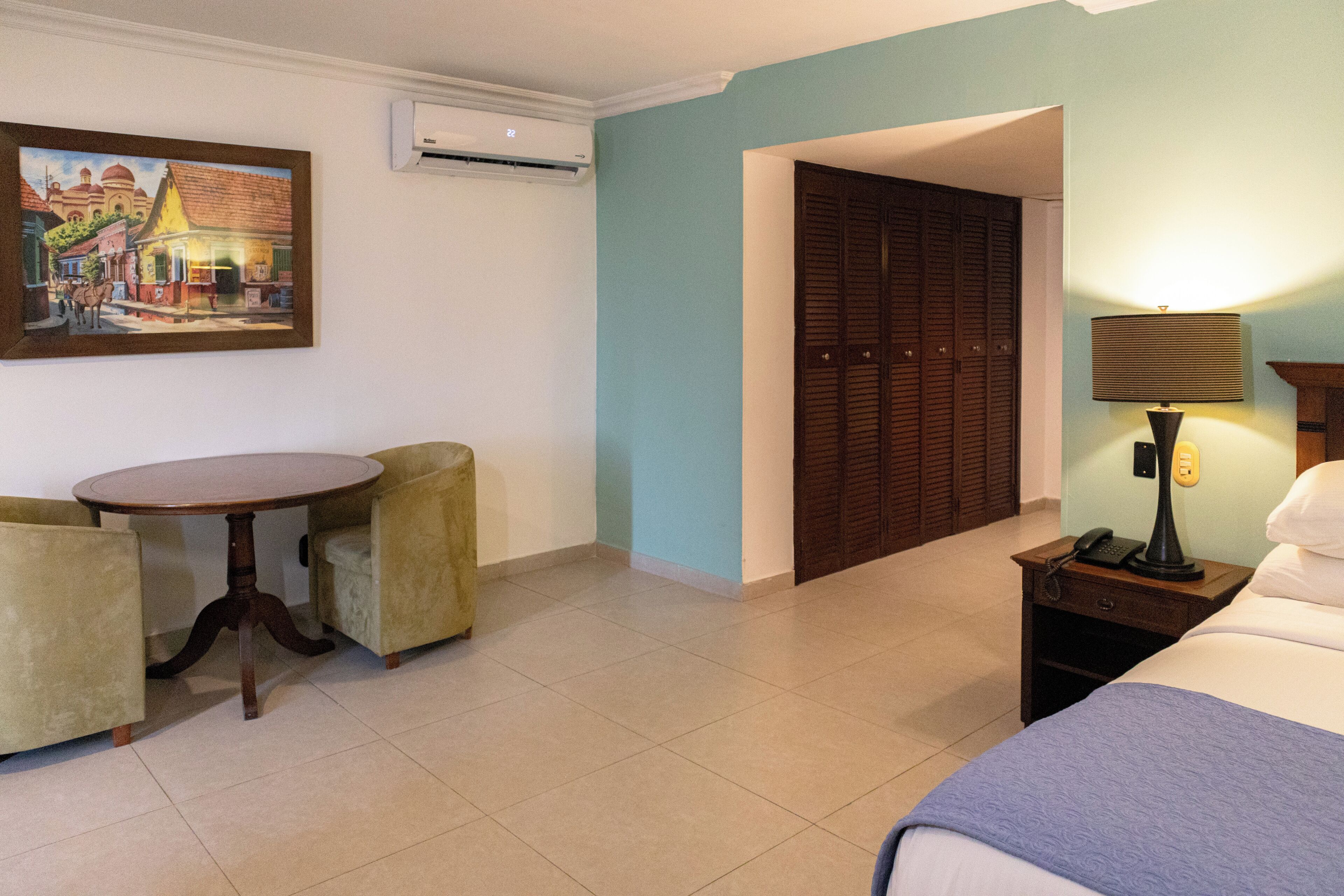 A hotel room with a bed, a table with two chairs, a painting on the wall, a wardrobe, and an air conditioning unit.