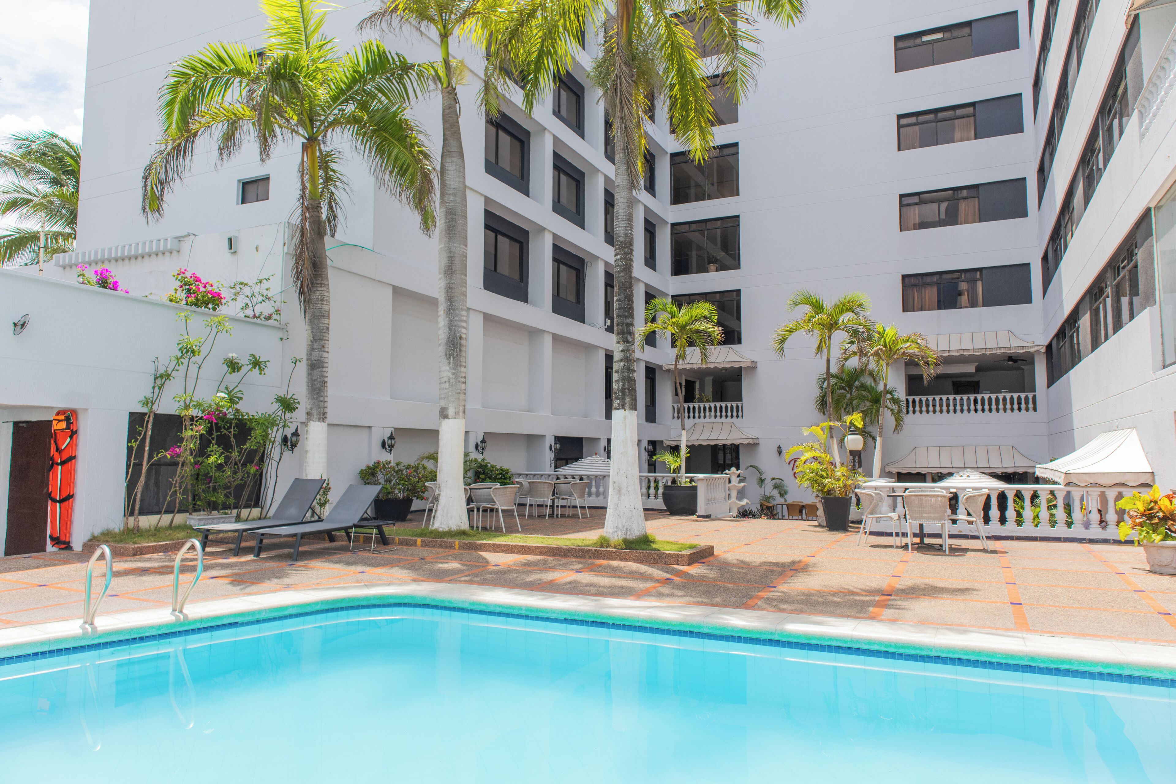 Área de piscina com espreguiçadeiras e palmeiras, cercada por um edifício com vários andares e varandas.