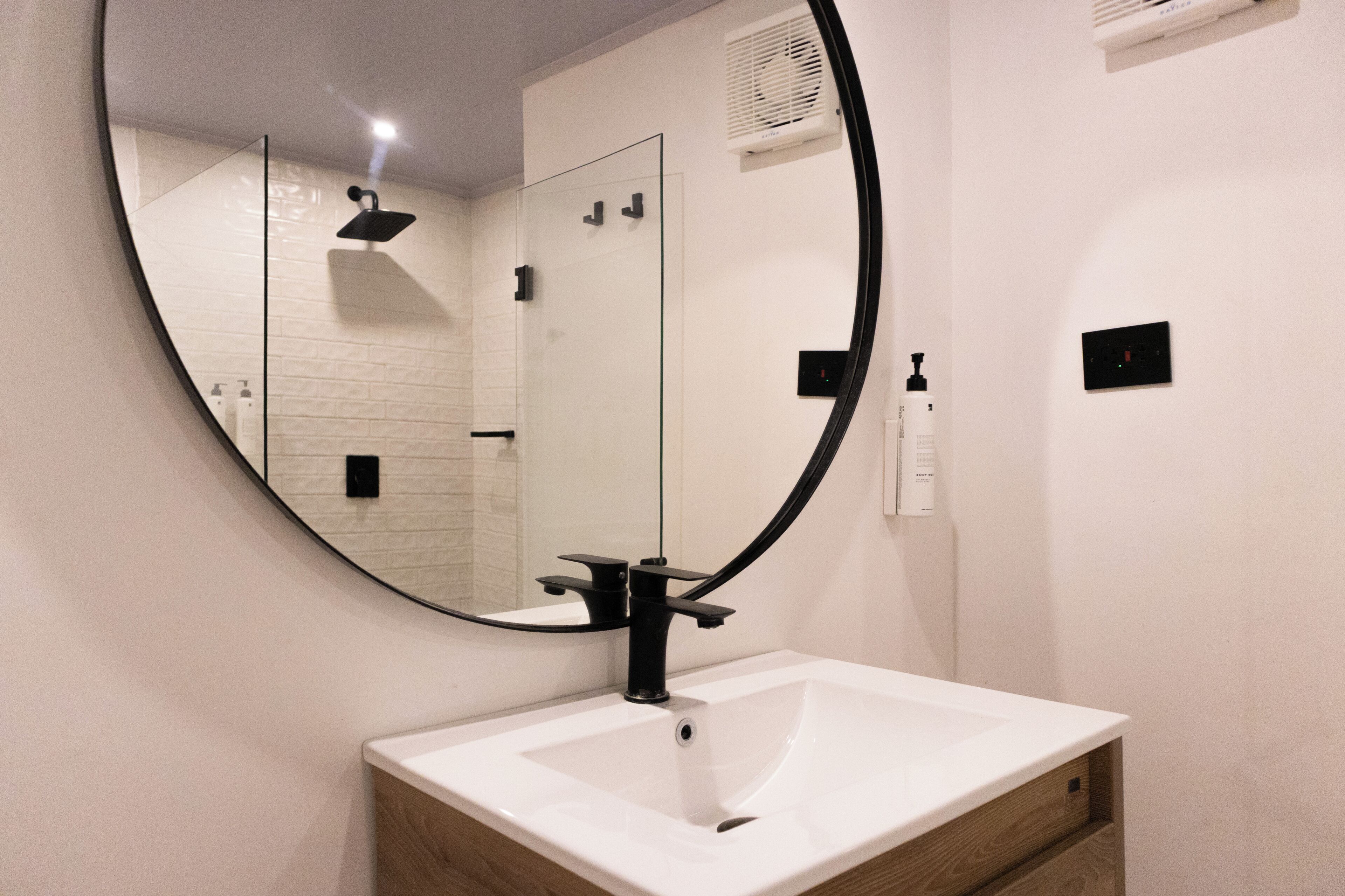 Un baño con un espejo redondo, un lavabo blanco y una mampara de vidrio para la ducha.