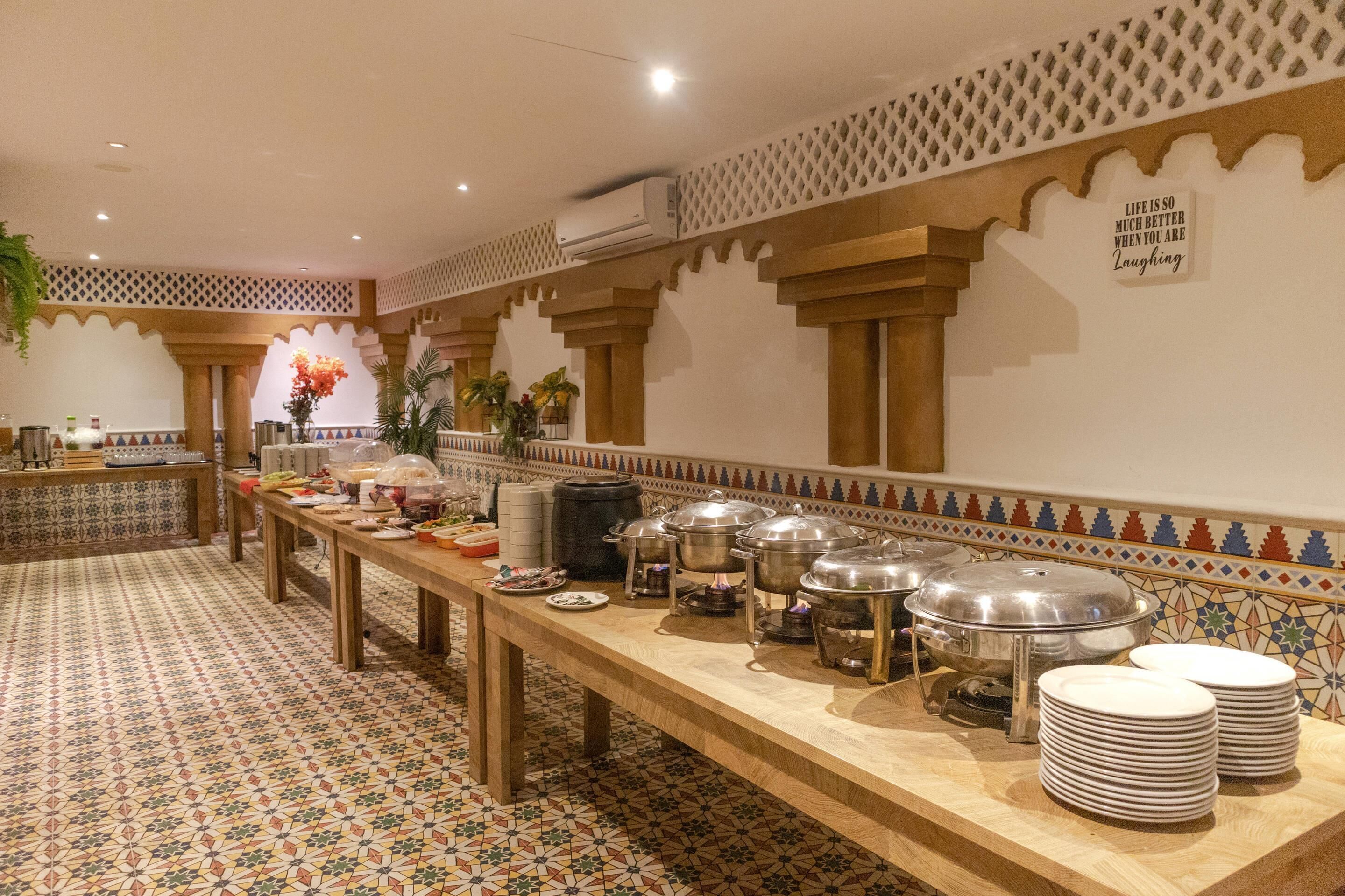 A buffet setup with a long table, food containers, and plates, set against a patterned wall with a decorative border.