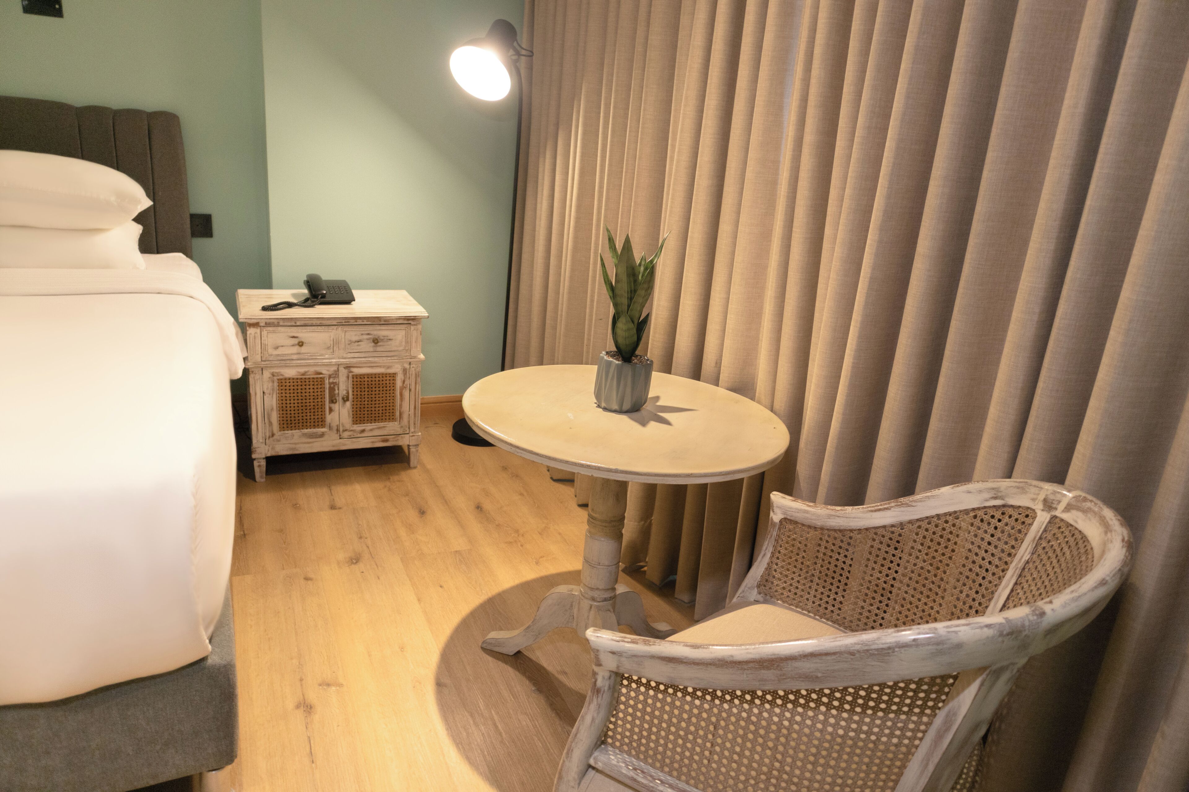Habitación de hotel con una cama, una mesita de noche con lámpara, una silla, una pequeña mesa con una planta y una ventana con cortinas.