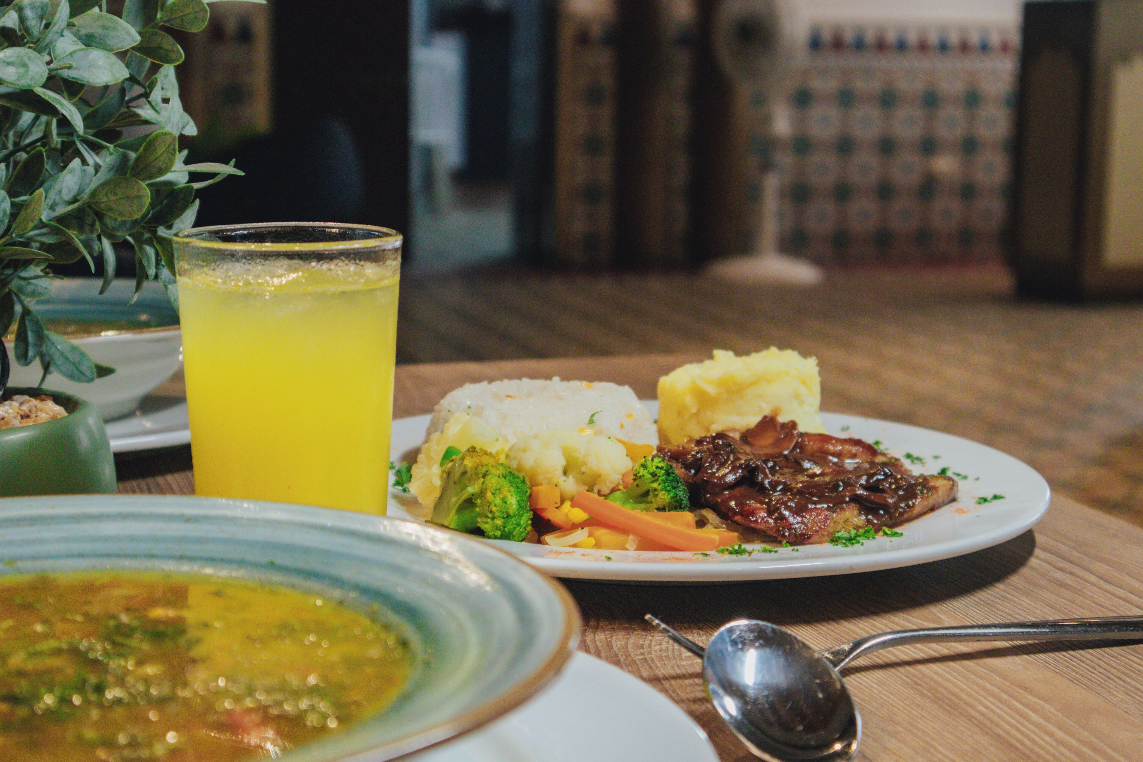 Una mesa de comedor con un plato de comida, un vaso de bebida amarilla y un tazón de sopa.
