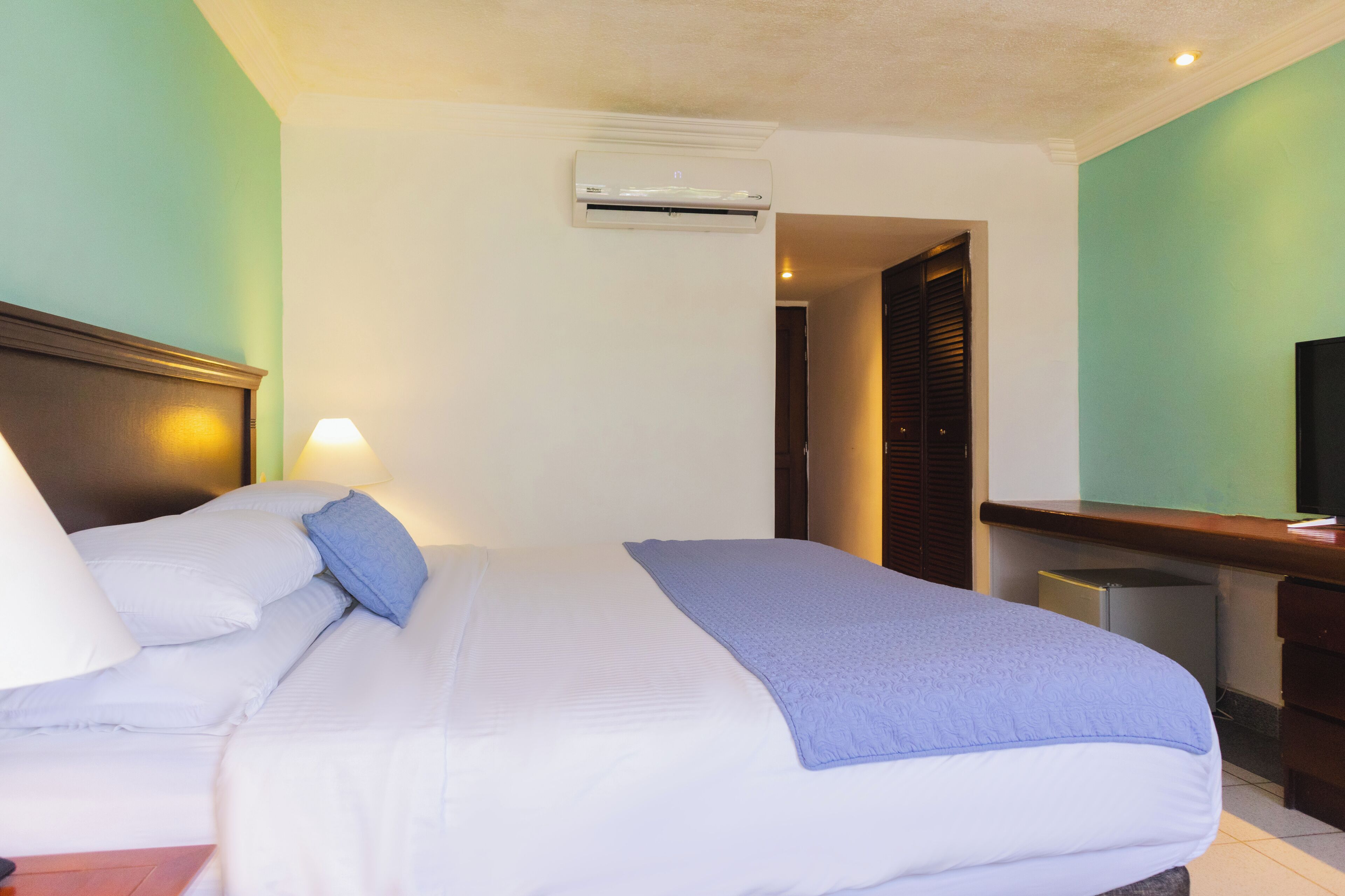 A hotel room with a bed, bedside lamps, an air conditioning unit, a desk, and a television.
