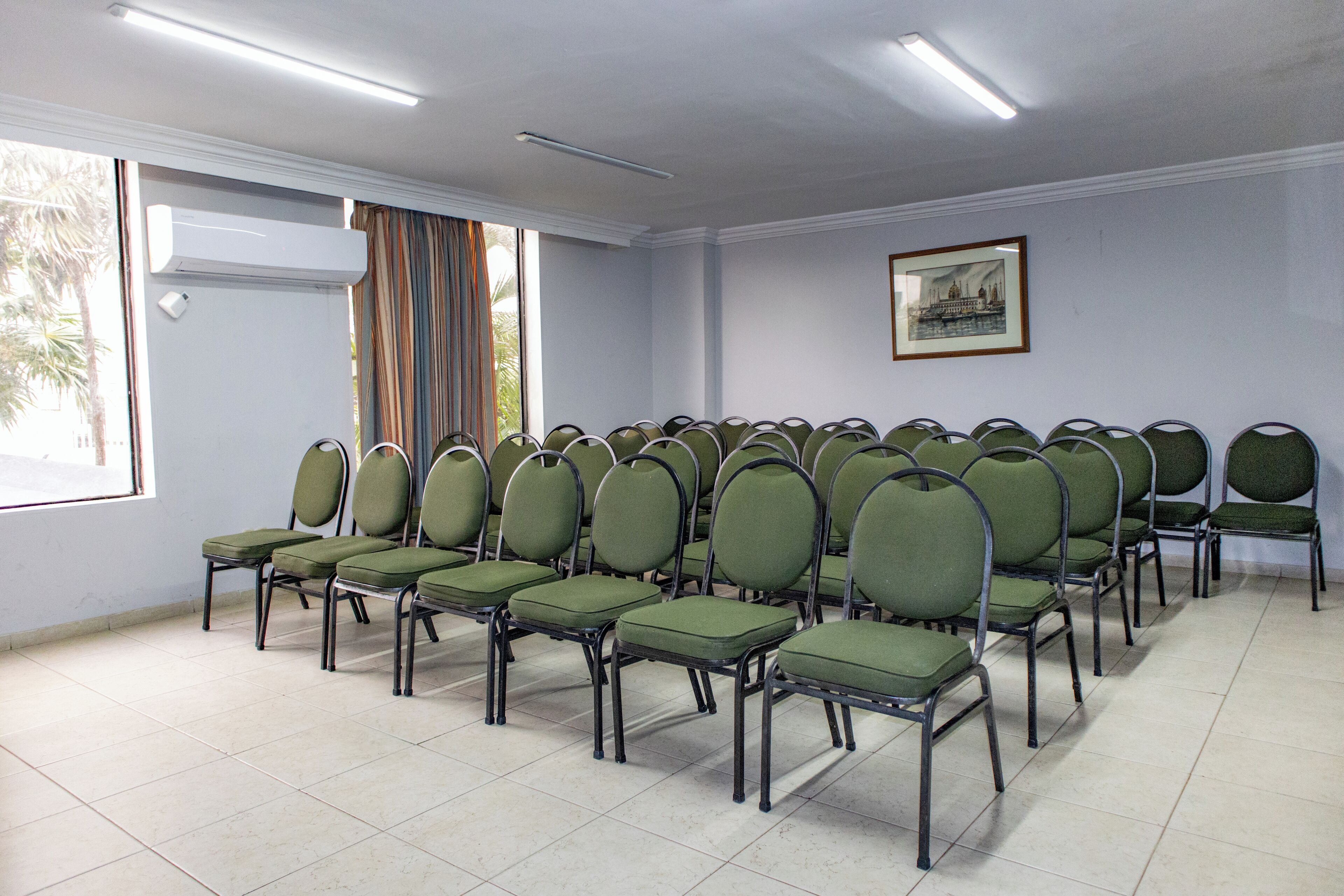 Sala de conferências com fileiras de cadeiras verdes, uma janela grande com cortinas e um quadro na parede.