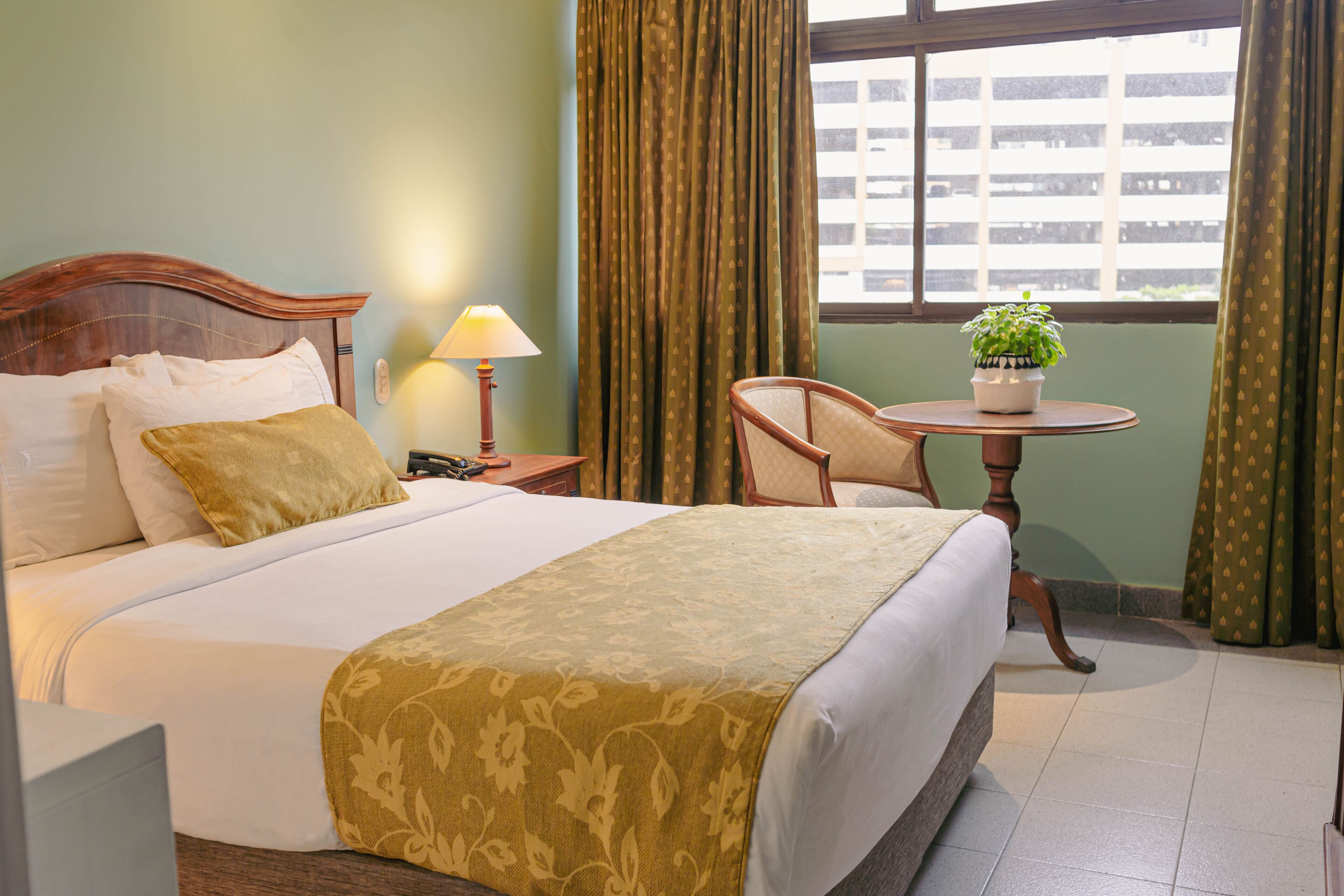 A hotel room with a large bed, a bedside table with a lamp, a chair, a small table with a plant, and a window with curtains.