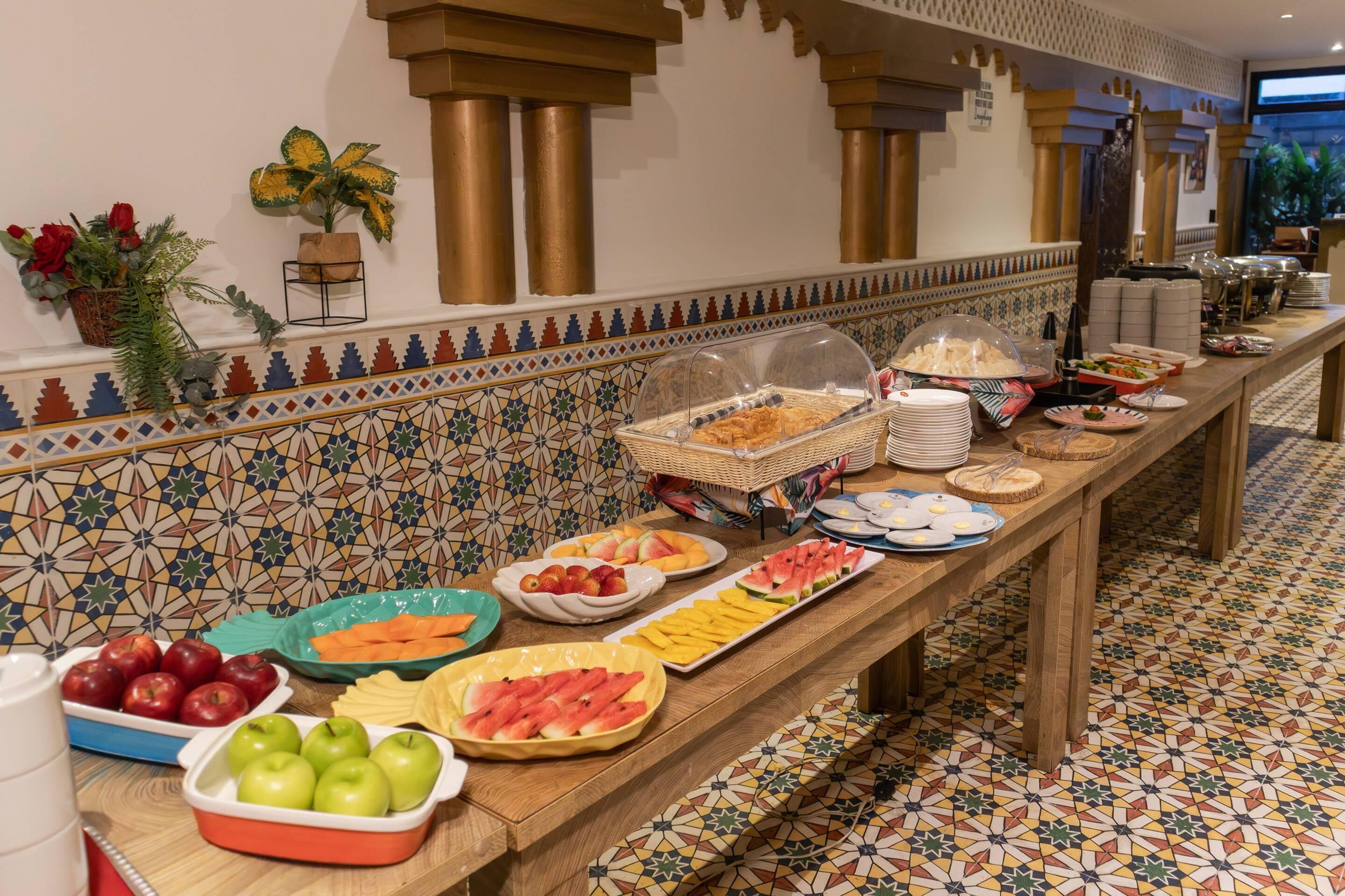 A buffet with various fruits and dishes, a tiled wall with a geometric pattern, and a wooden floor.
