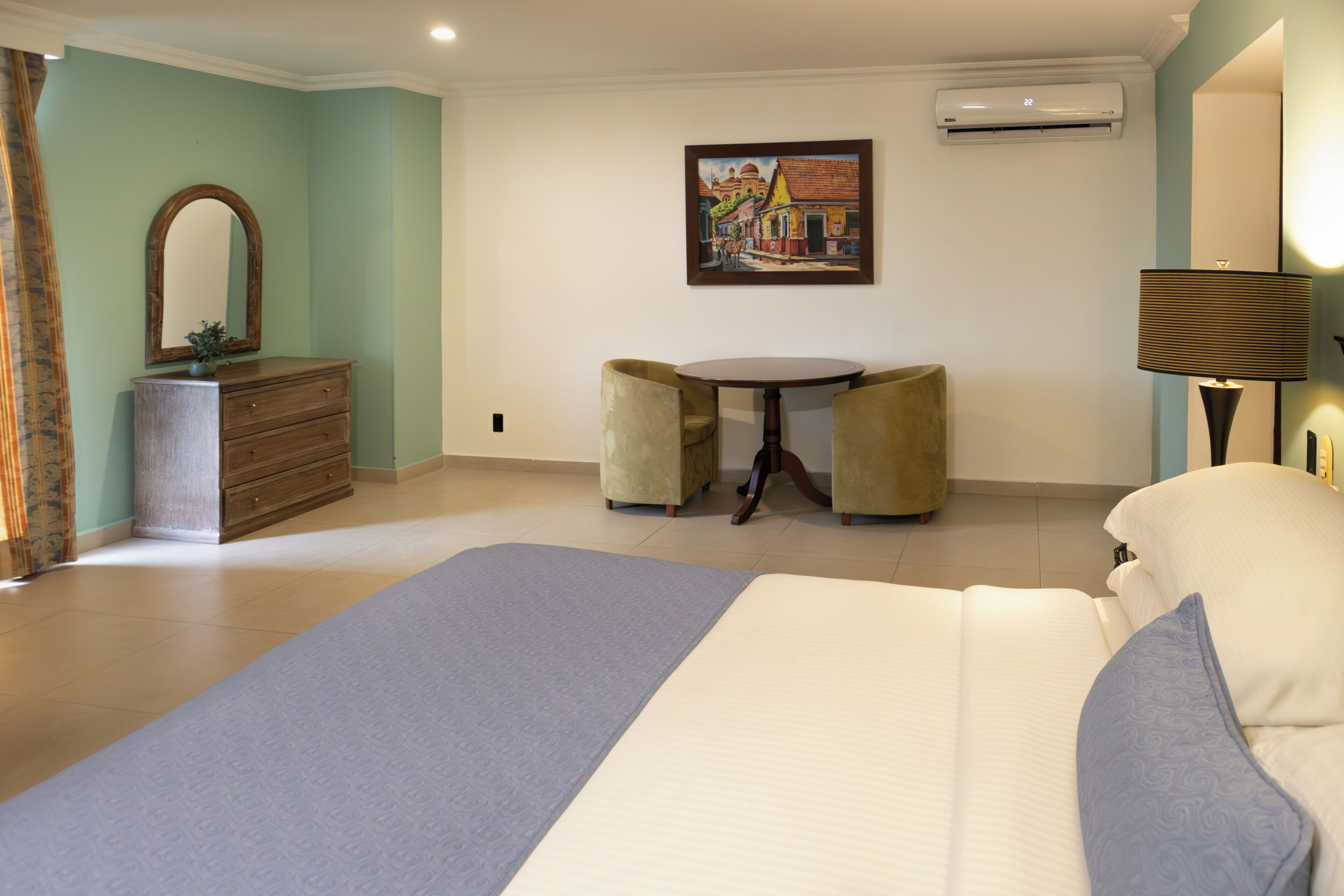 A hotel room with a bed, a dresser, a round table with chairs, a painting on the wall, and an air conditioning unit.