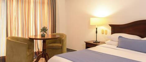 A hotel room with a bed, a round table with a plant, two armchairs, a nightstand with a lamp, and curtains.
