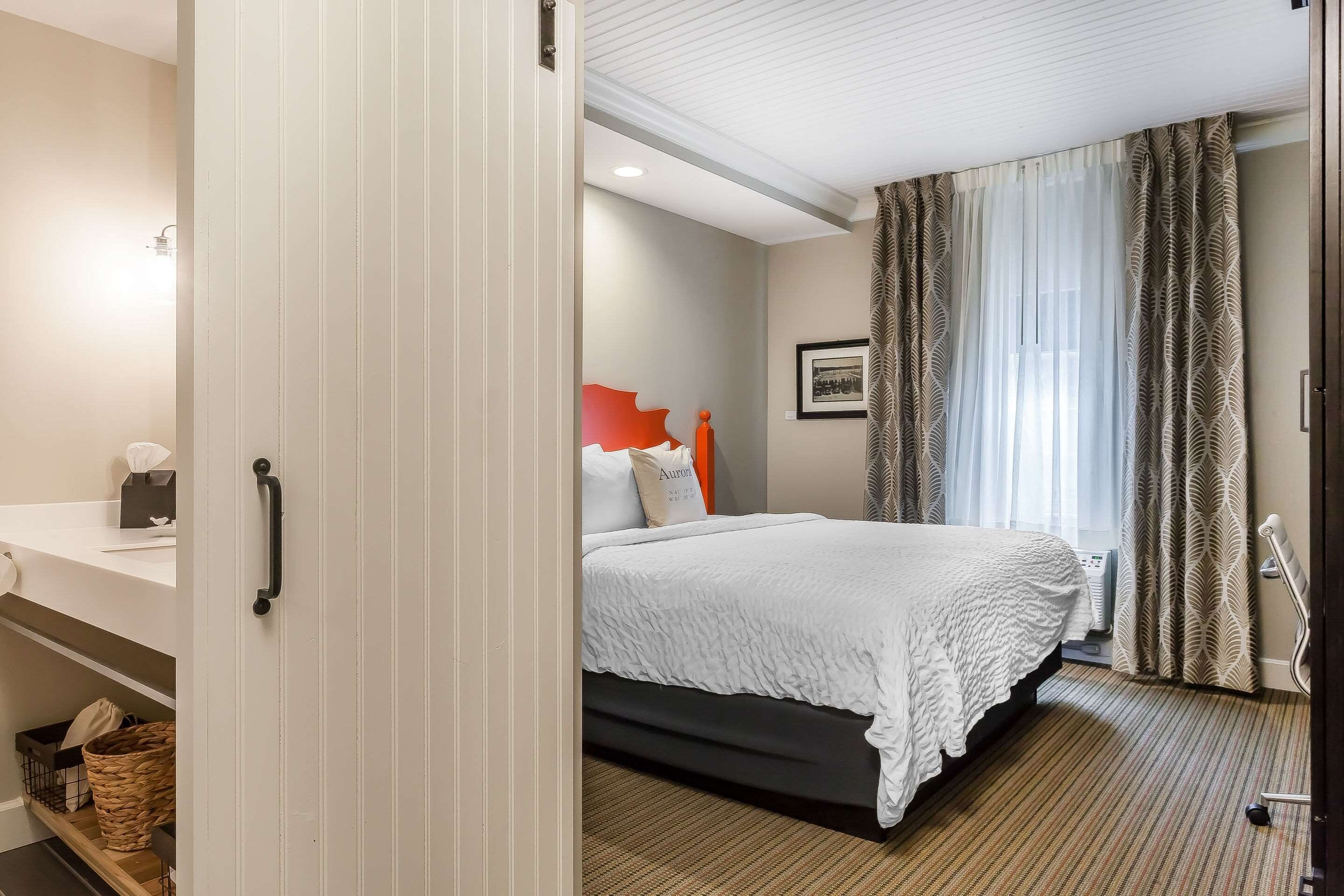 A hotel room with a bed, a bedside table, a shelf with baskets, a framed picture on the wall, and a window with curtains.