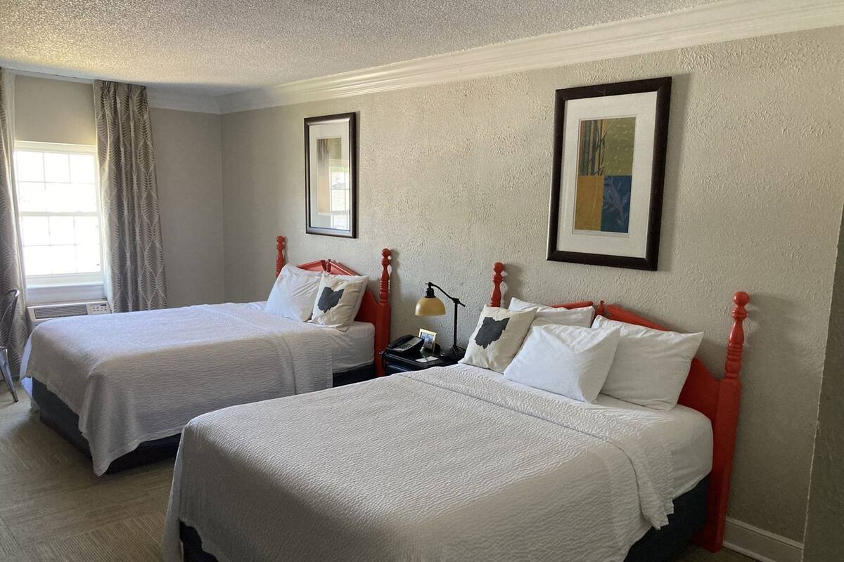 A hotel room with two beds, a nightstand, a lamp, and a window with curtains.