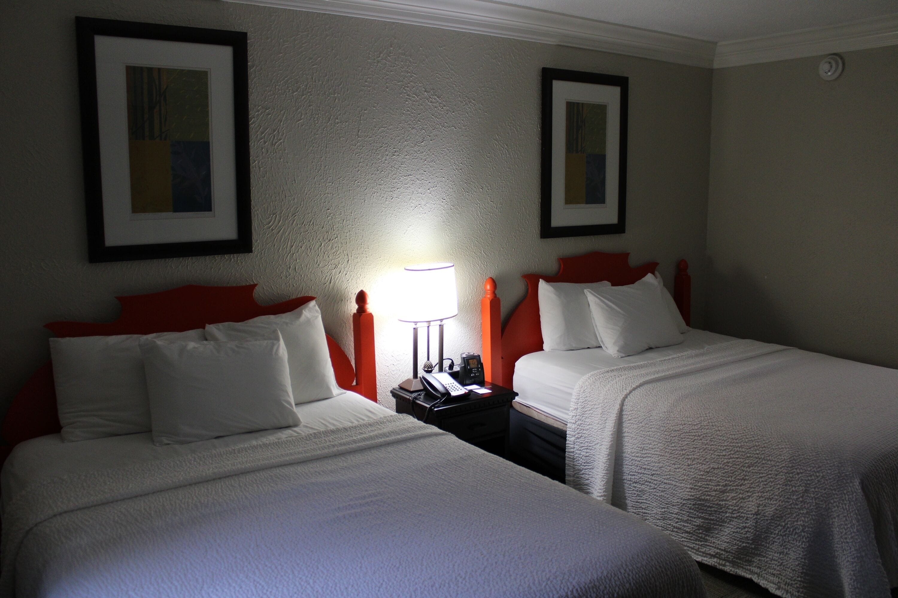 Habitación de hotel con dos camas, cada una con ropa blanca y cabeceros rojos. Hay una mesita de noche con una lámpara y un teléfono entre las camas.