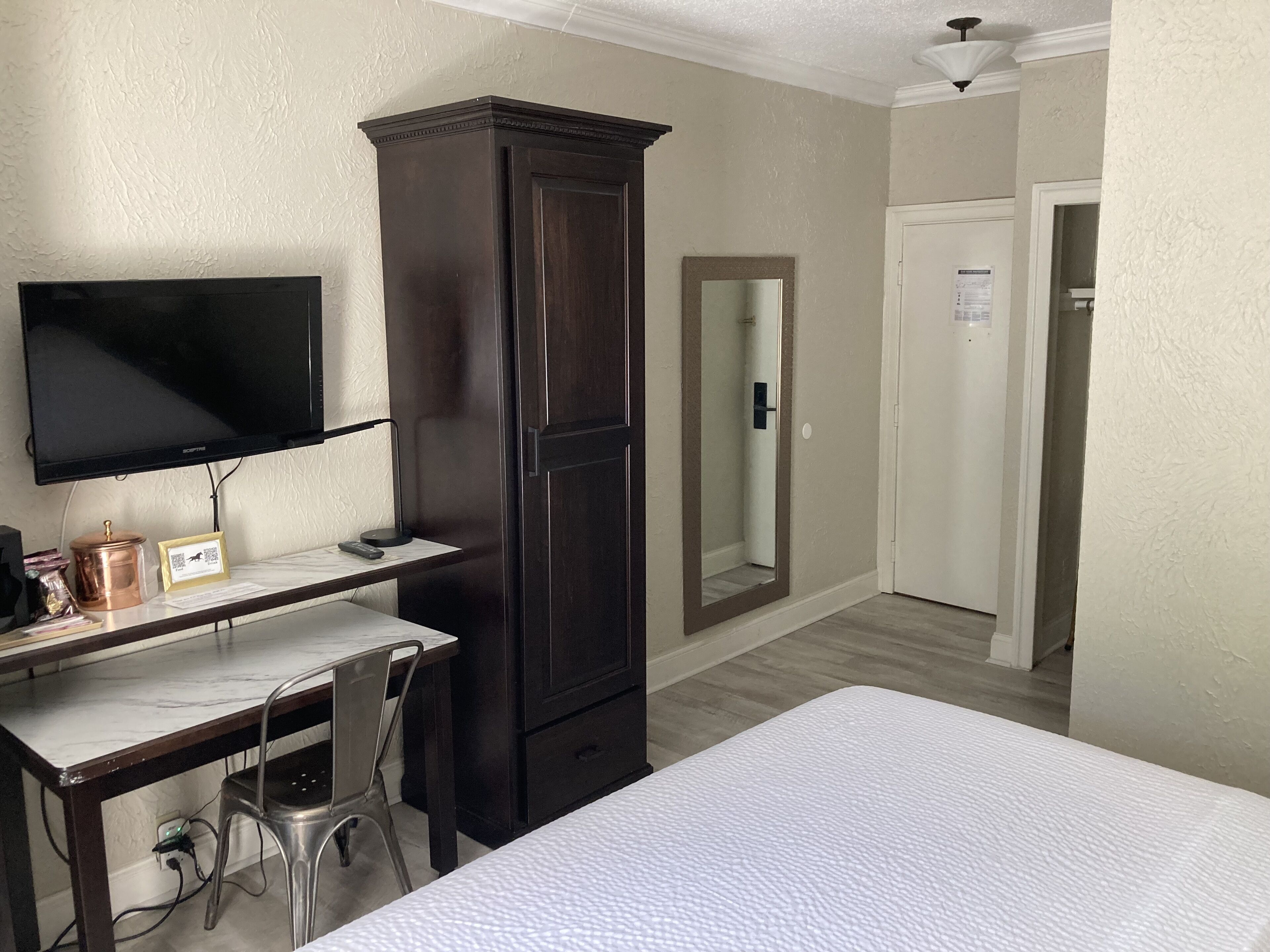 Habitación de hotel con cama, escritorio con silla, armario, televisor y puerta que da a otra habitación.