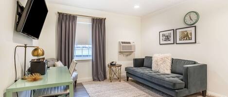 A modern living room with a glass desk, a grey sofa, a mounted TV, a clock, and framed pictures on the wall.