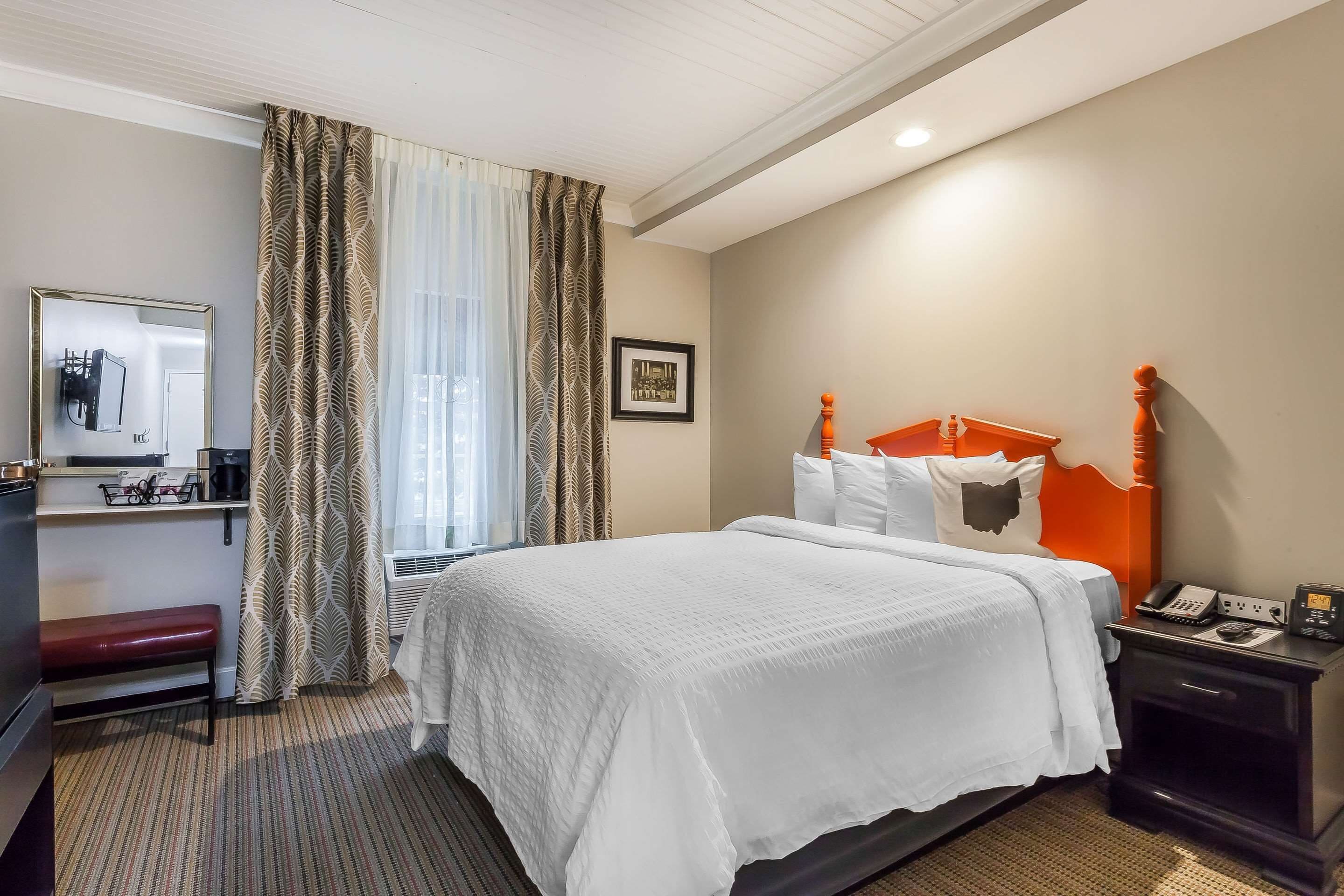 A hotel room with a large bed, a bedside table with a phone and a lamp, a bench, a mirror, and a picture on the wall.