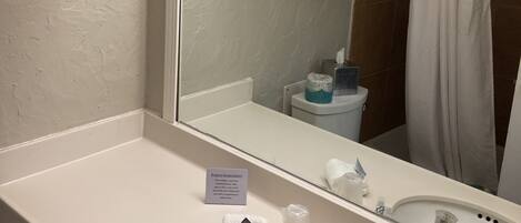 A bathroom with a large mirror, two sinks, a towel rack with folded towels, a toilet paper holder, and a sign offering assistance for forgotten items.
