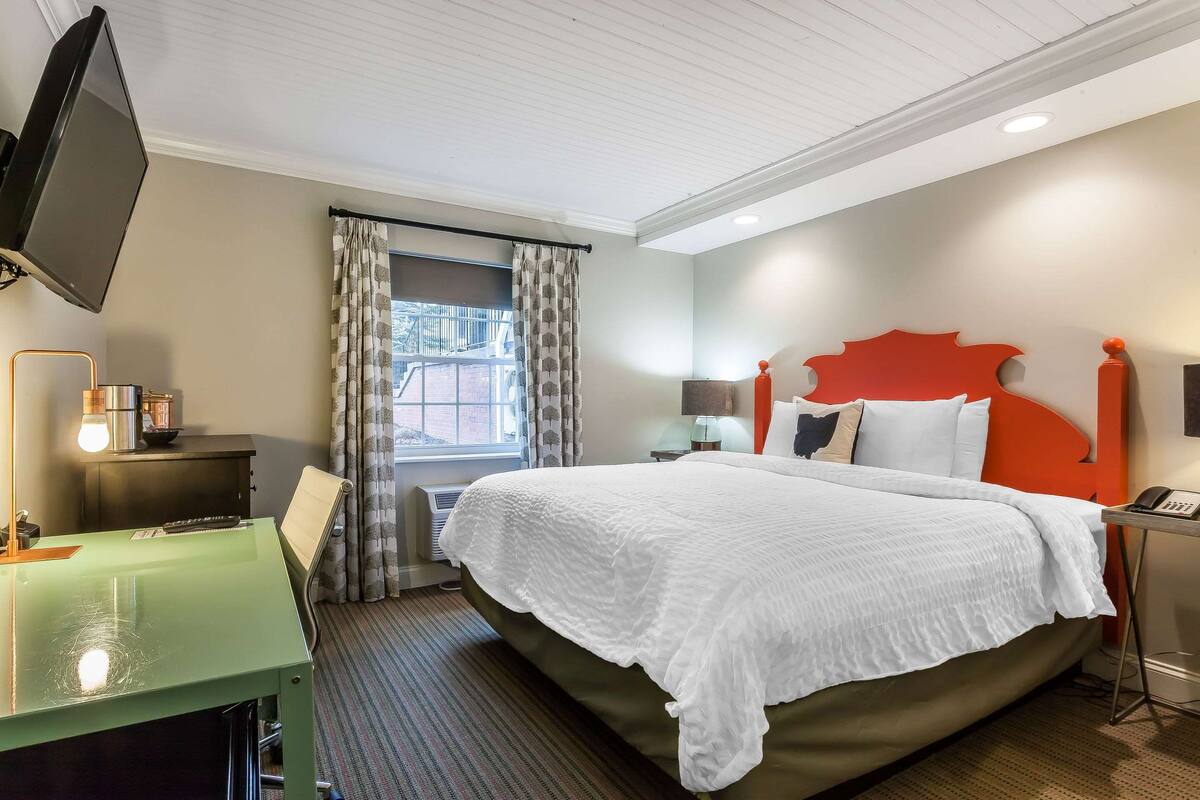A hotel room with a bed, a desk with a lamp, a TV, and a window with curtains.