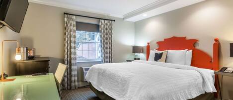 A hotel room with a bed, a desk with a lamp, a TV, and a window with curtains.