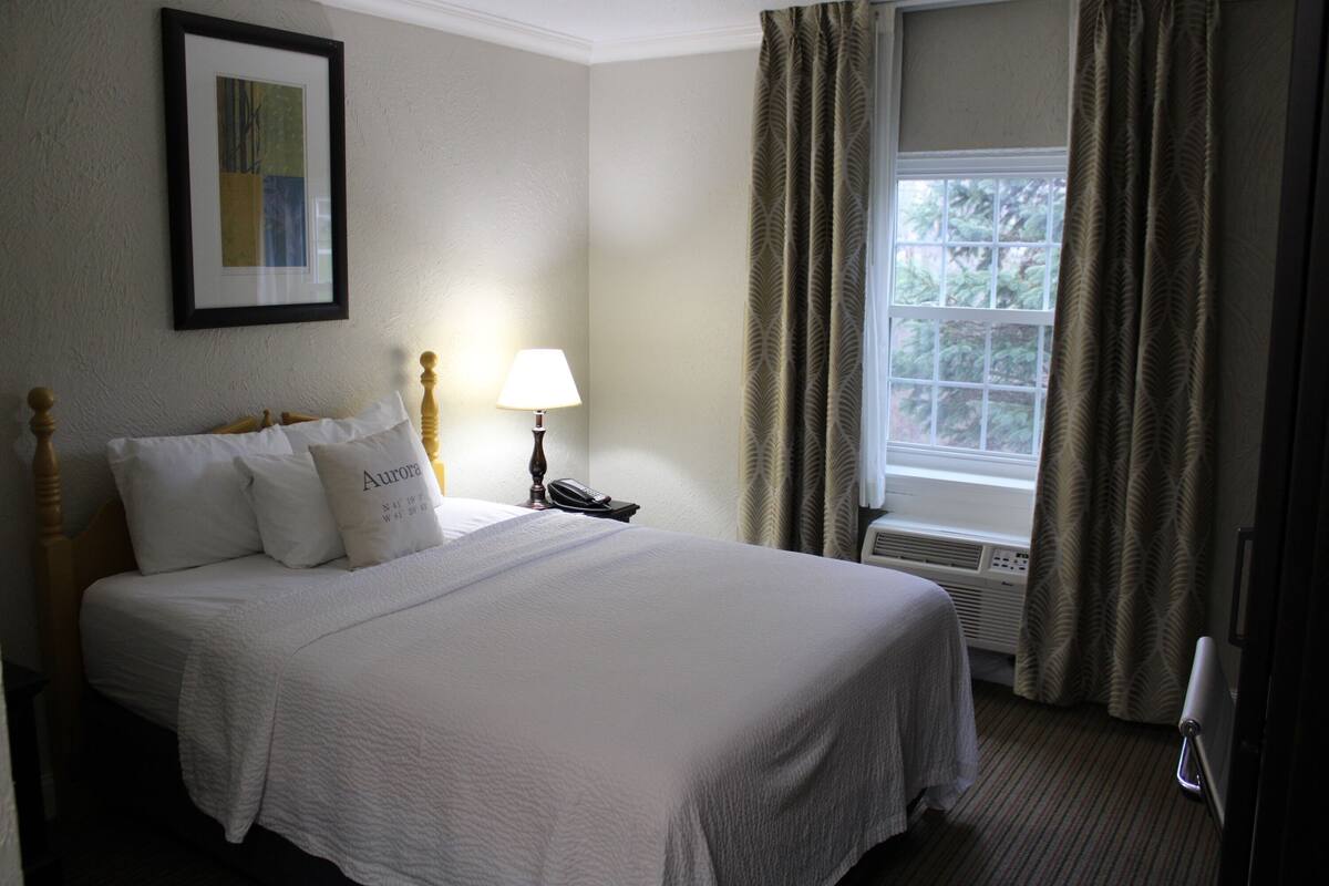 A neatly made bed with white bedding and pillows, a bedside lamp, a framed picture on the wall, and a window with curtains.