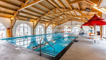 An indoor swimming pool with a high wooden ceiling, large windows, and a red umbrella.