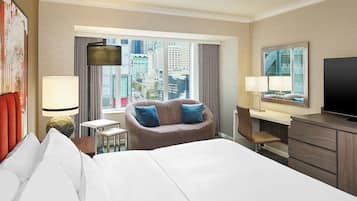 A hotel room with a large bed, a desk, a chair, a TV, and a view of the city.