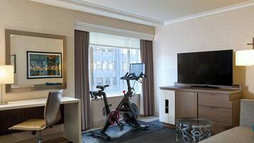 Habitación de hotel con escritorio, silla, televisión, bicicleta estática y ventana con cortinas.