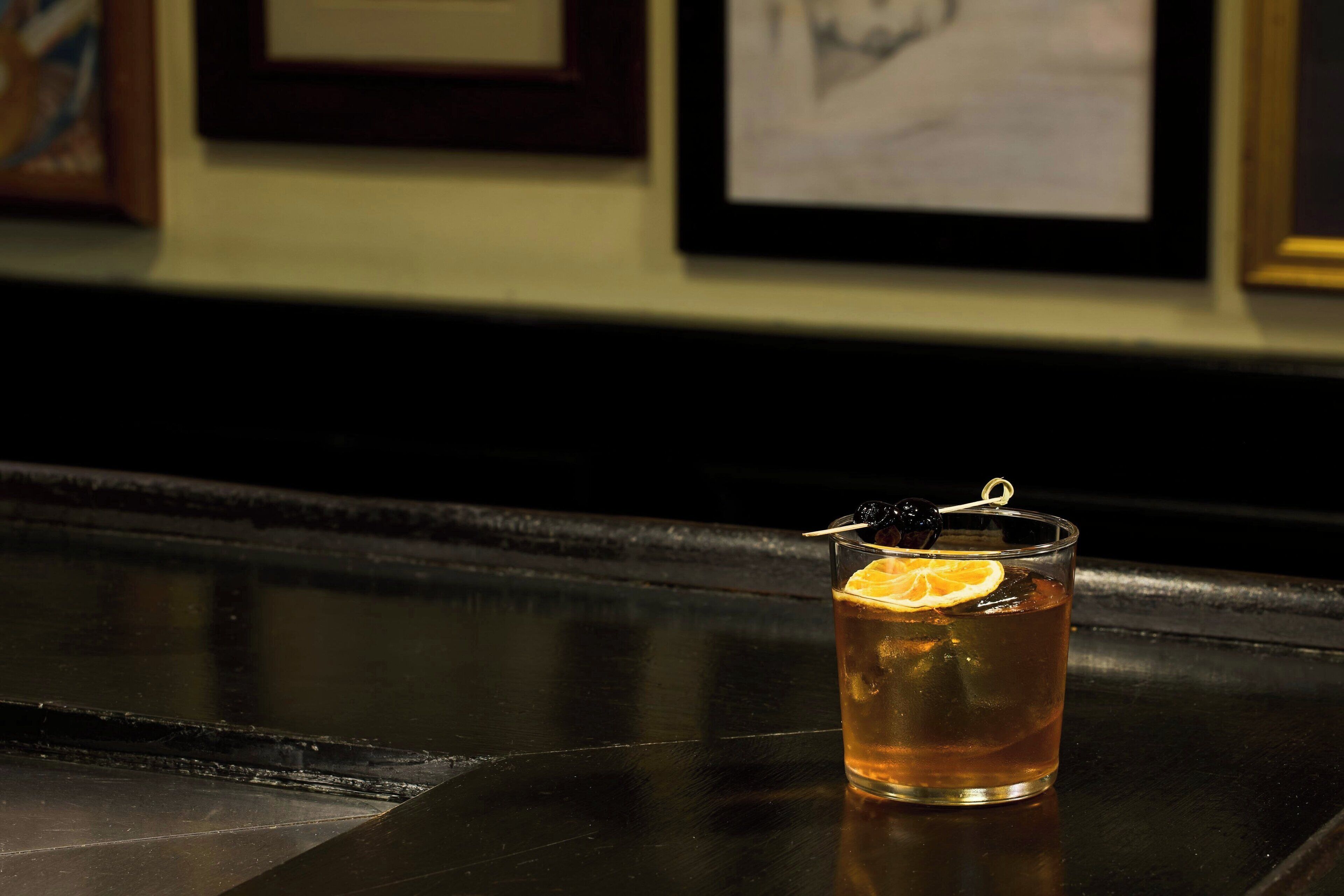Un verre contenant une boisson de couleur ambrée, orné d’une tranche d’orange et d’un fruit noir sur le bord, posé sur une surface réfléchissante.