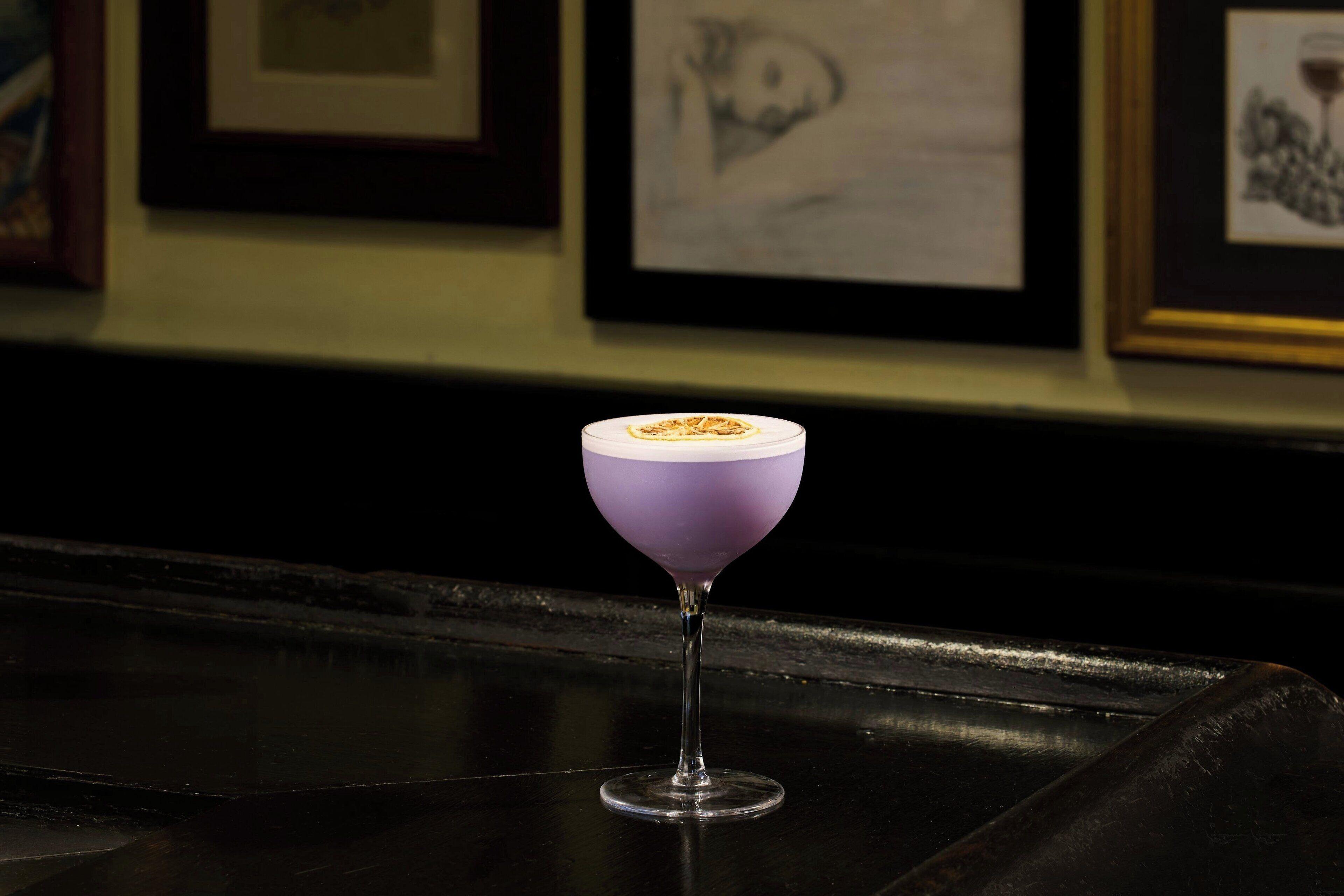 Un bar avec une boisson violette servie dans un verre, des œuvres d’art encadrées accrochées au mur et un comptoir noir.