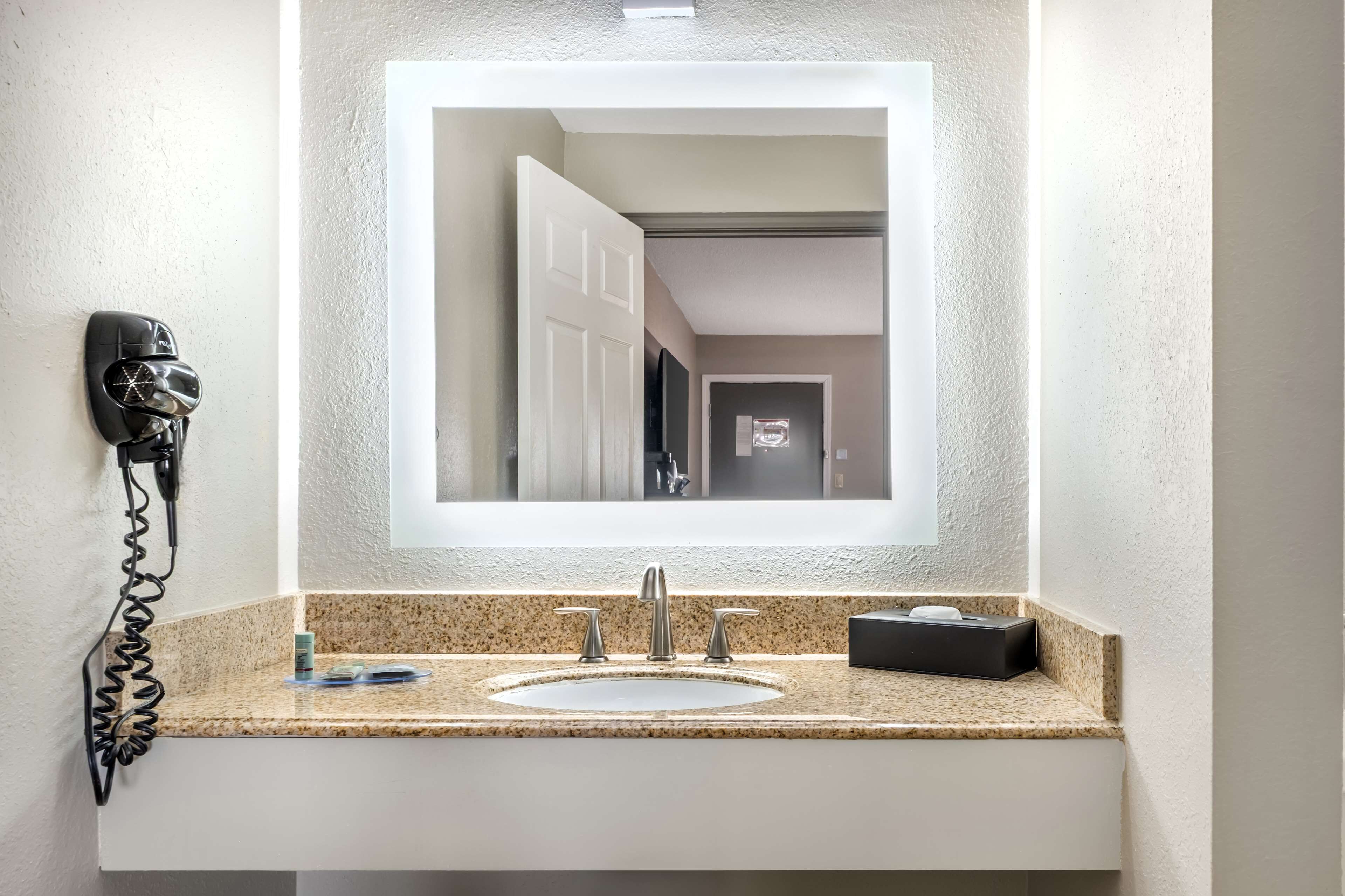 Un baño con encimera de granito, un lavabo blanco, un espejo, un secador de pelo y una caja pequeña sobre la encimera.