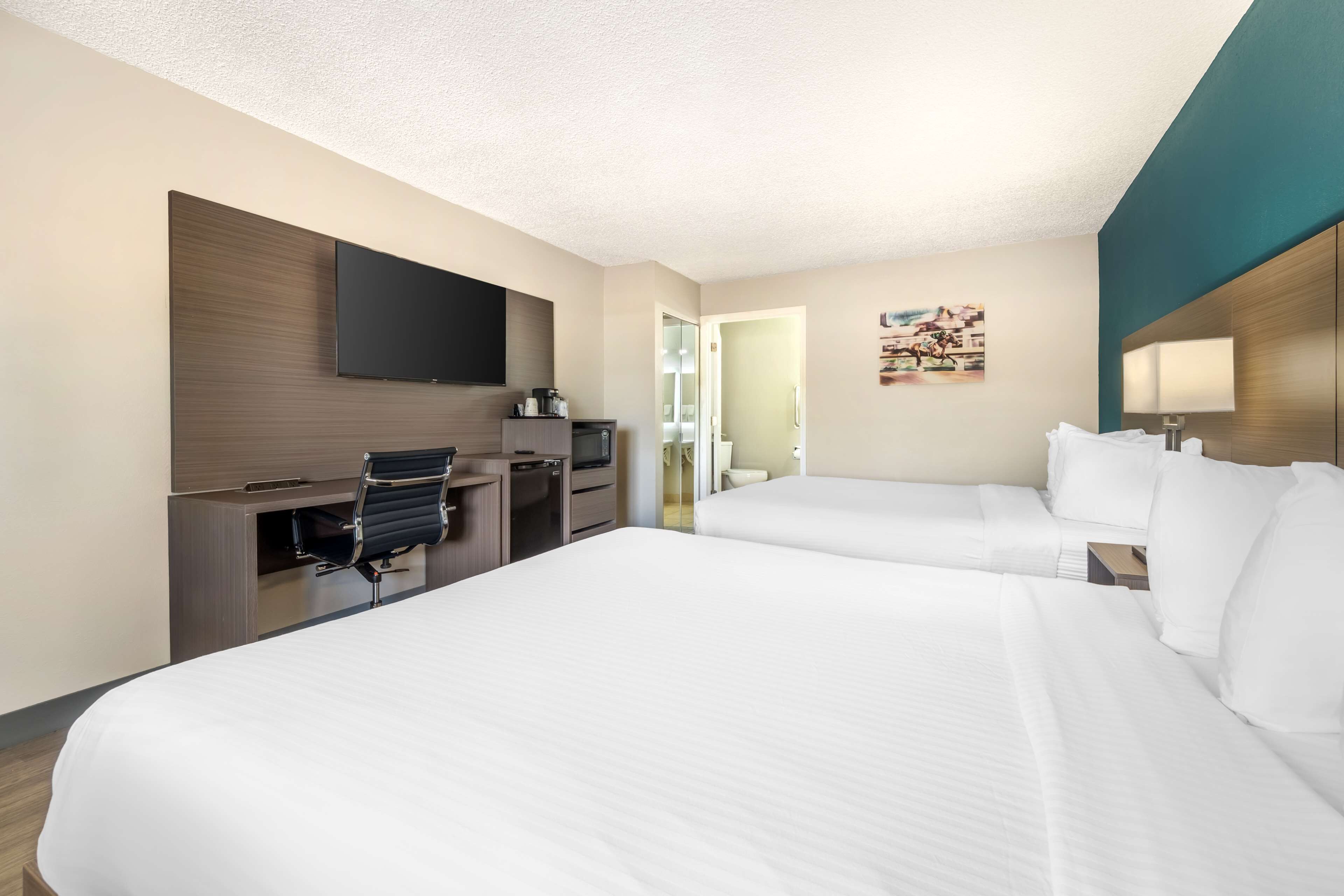 A hotel room with two beds, a desk with a chair, a flat-screen TV, and a small kitchenette.