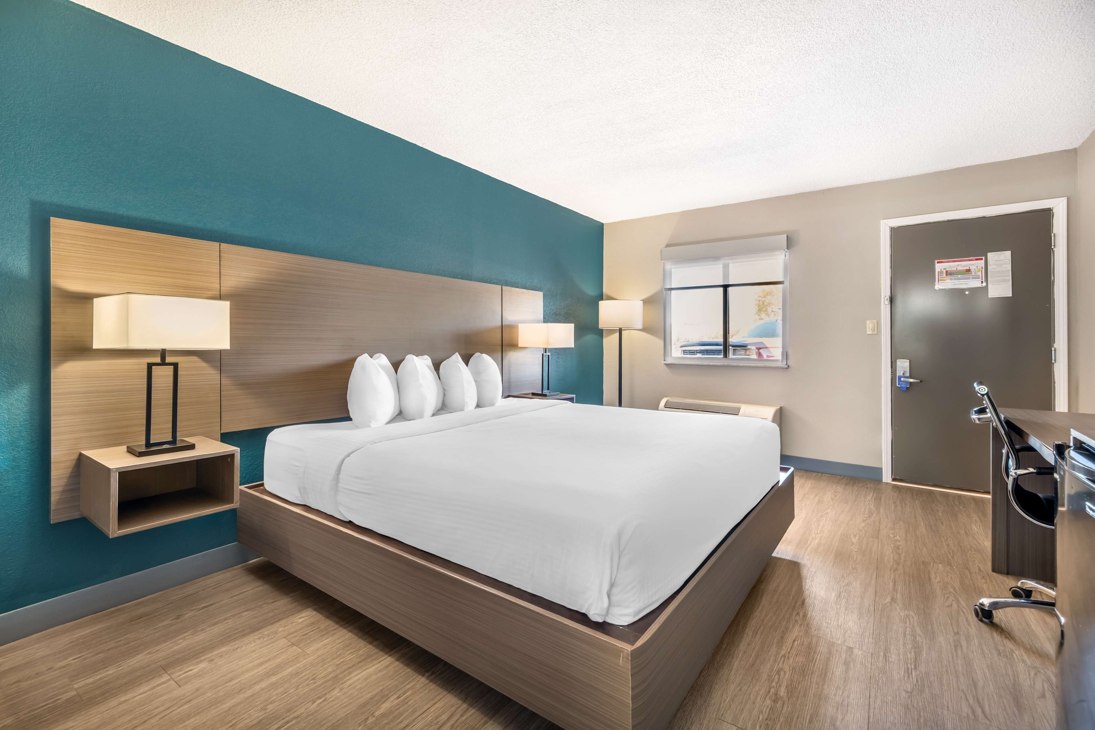 A modern hotel room with a large bed, two bedside tables with lamps, a wooden headboard, and a door with a bulletin board.