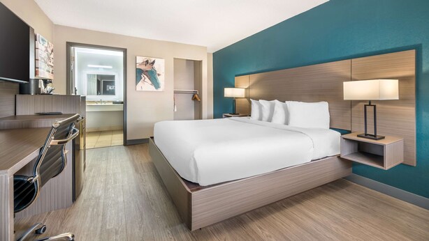 A modern hotel room with a large bed, a desk with a chair, a nightstand with a lamp, and artwork on the walls.