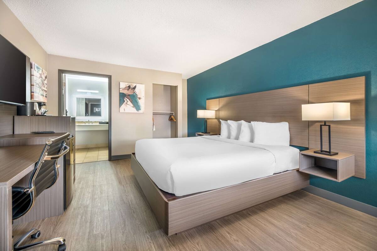 A modern hotel room with a large bed, a desk with a chair, a nightstand with a lamp, and artwork on the walls.