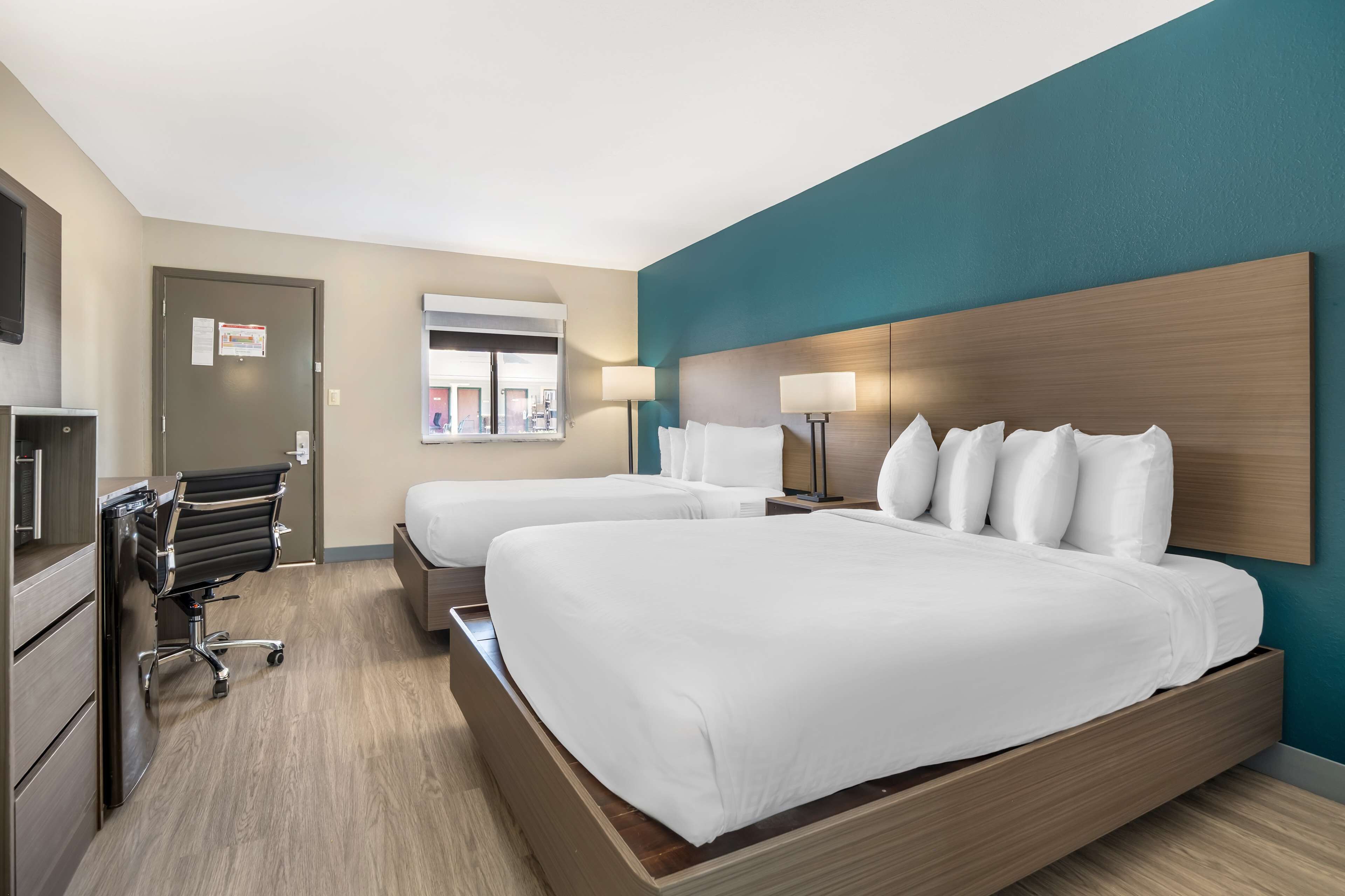 A hotel room with two beds, a desk, a chair, and a view of the outside.