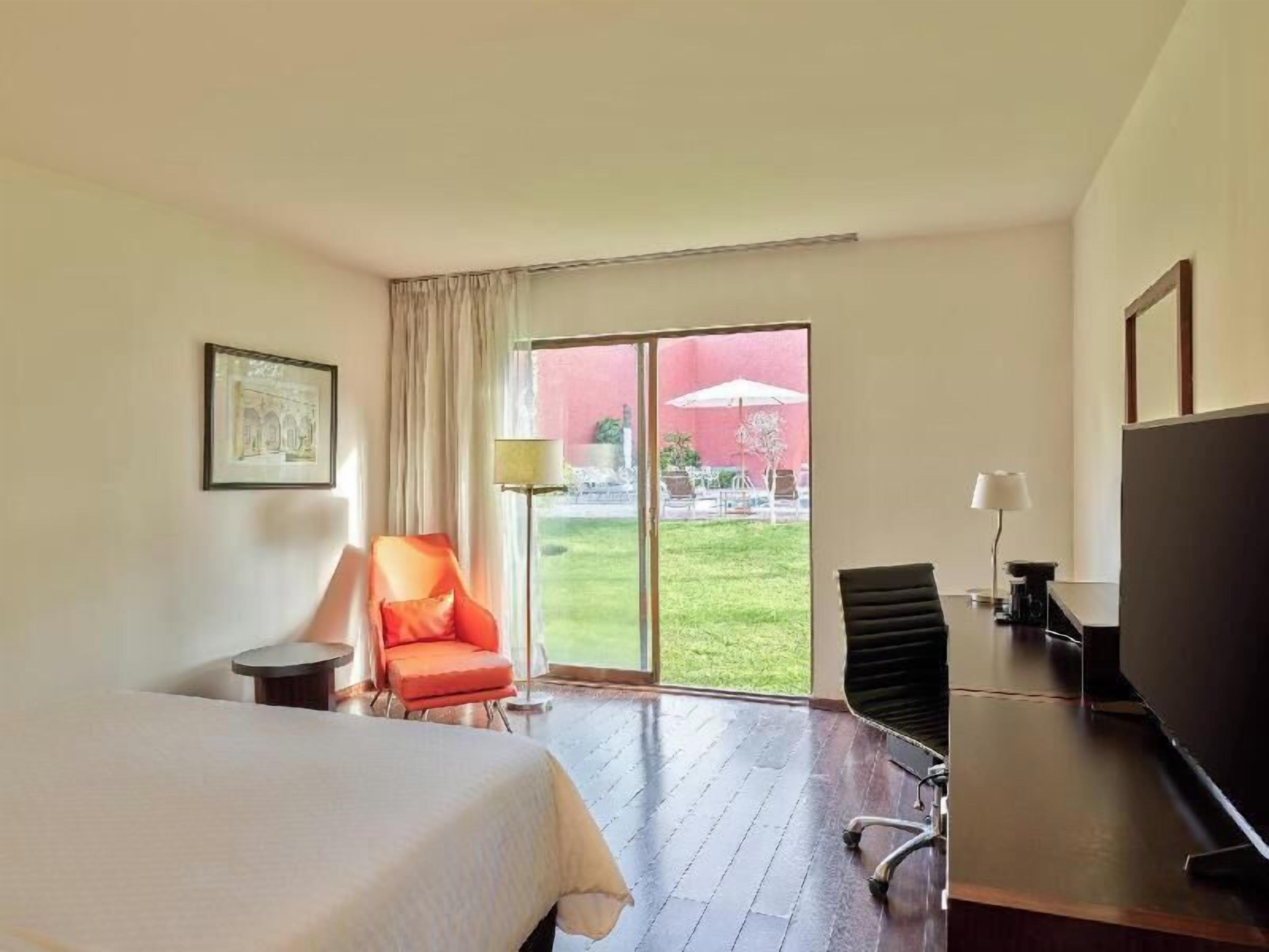 Habitación de hotel con cama, escritorio, silla, televisor y un amplio ventanal corredero que da a un área exterior.