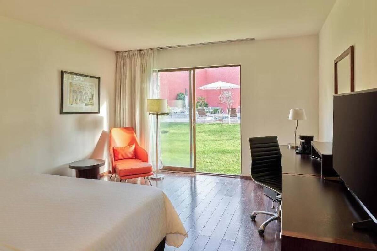 Quarto de hotel com cama, escrivaninha, cadeira, televisão e uma grande porta de vidro deslizante que dá para uma área externa.