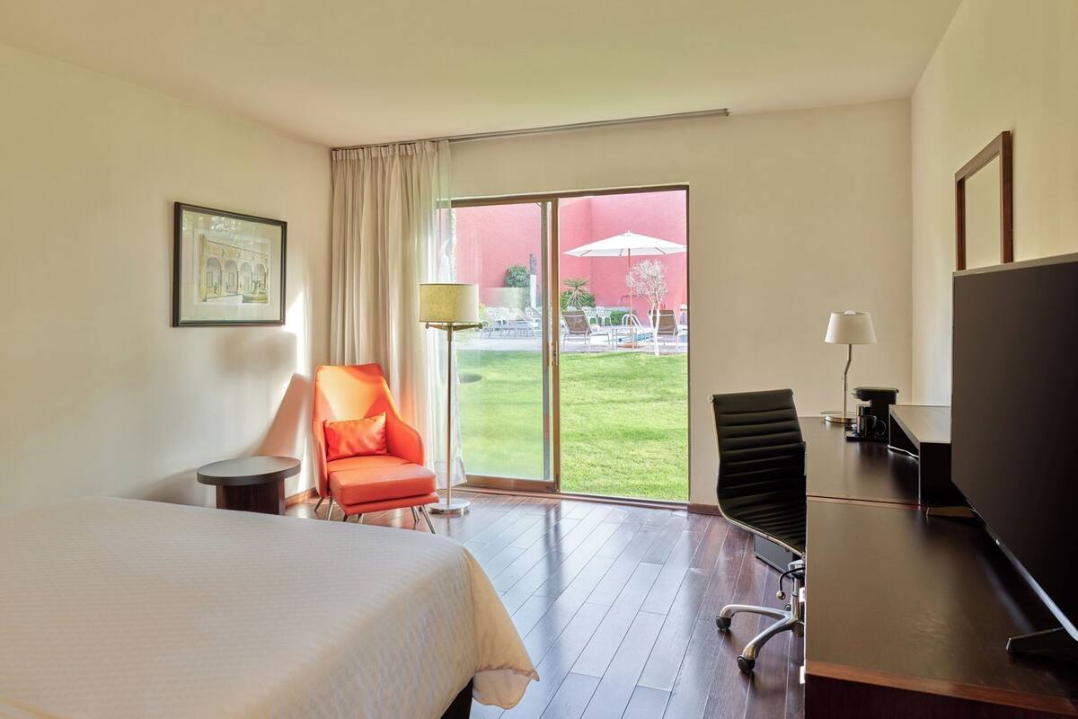 Quarto de hotel com cama, escrivaninha, cadeira, televisão e uma grande porta de vidro deslizante para área externa.