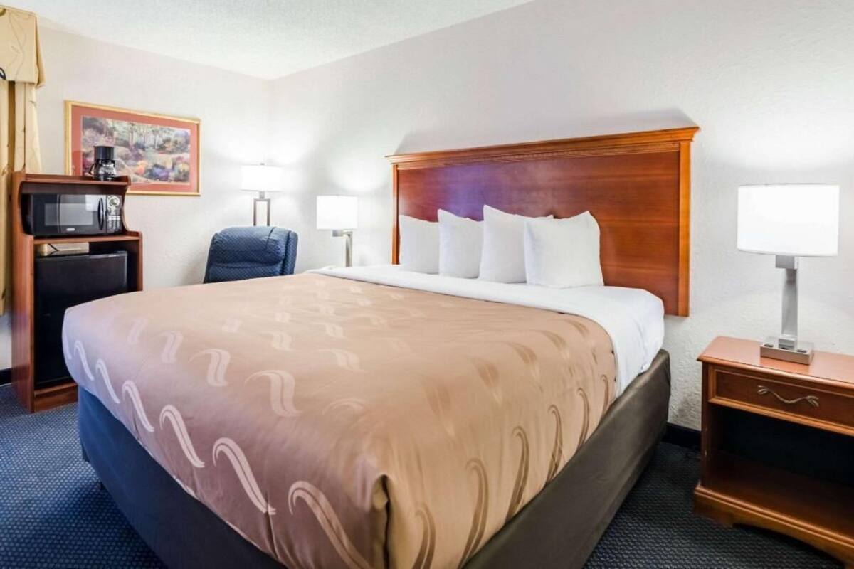 A hotel room with a large bed, a desk with a computer, a chair, a lamp, and a framed picture on the wall.