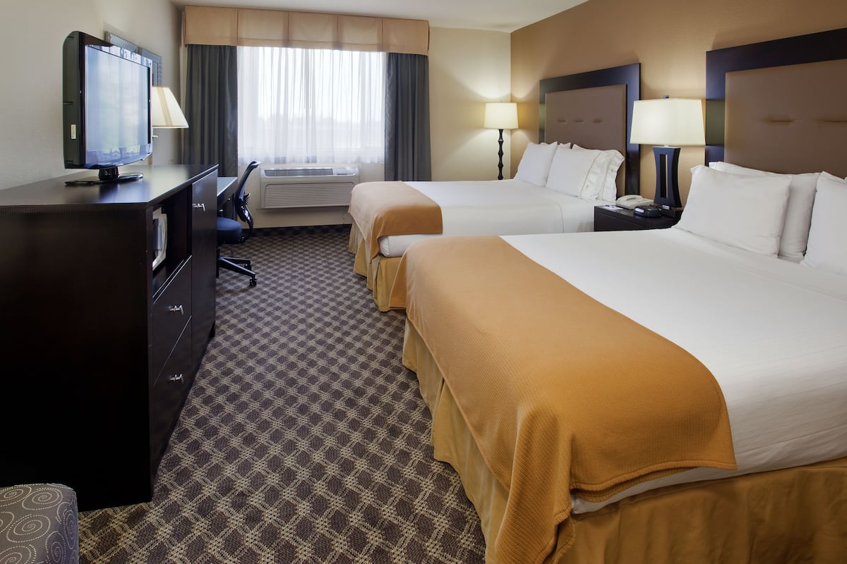 Quarto de hotel com duas camas, uma mesa de trabalho e uma televisão, além de uma janela com cortinas.