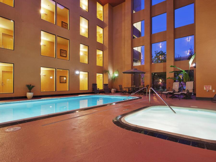 An indoor swimming pool with multiple lanes, surrounded by lounge chairs and tables, and a large building with many windows in the background.