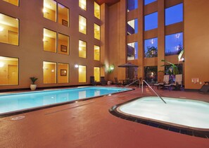 An indoor swimming pool with multiple lanes, surrounded by lounge chairs and tables, and a large building with many windows in the background. - Holiday Inn Express Union City (San Jose) by IHG (Union City)