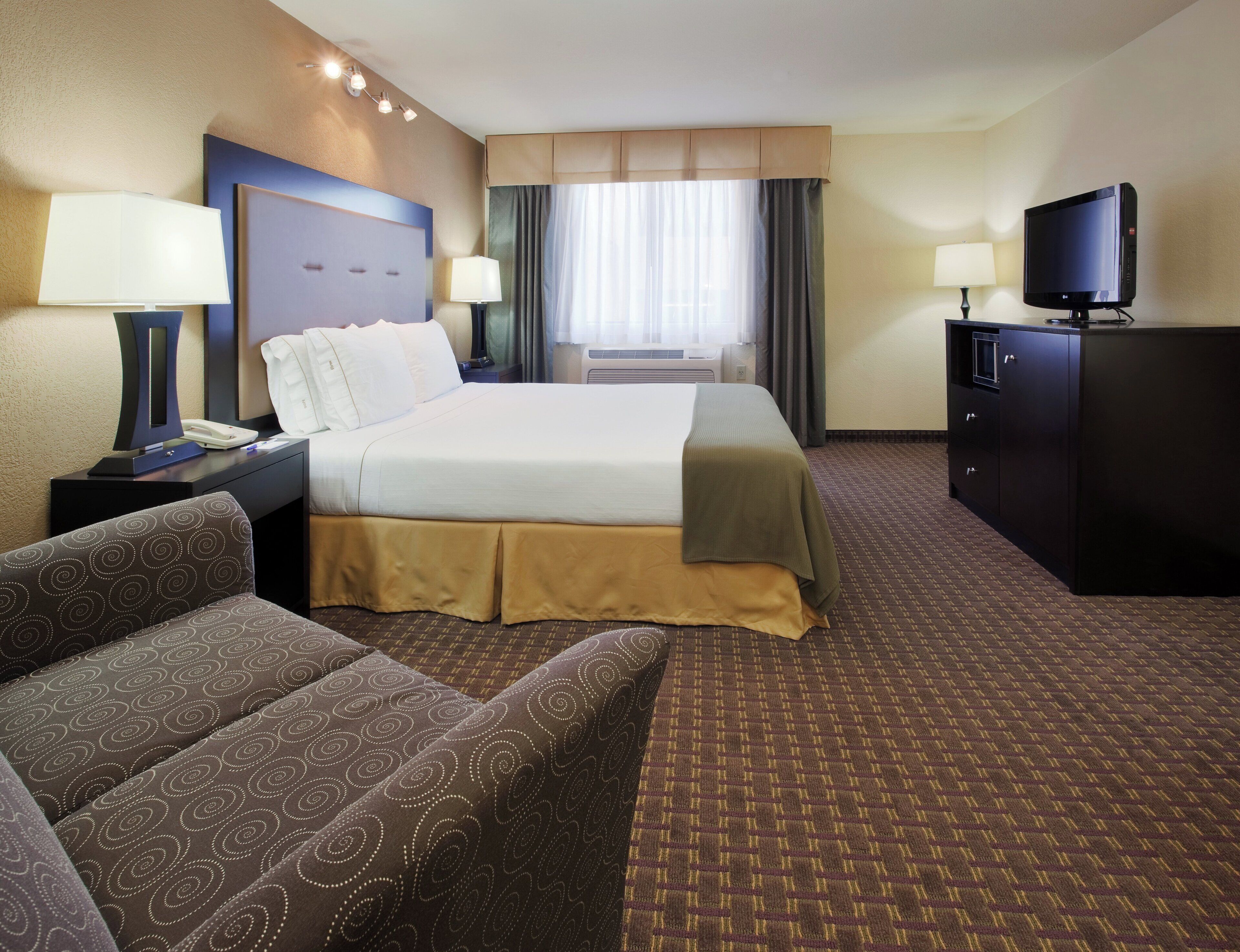 A hotel room with a bed, two bedside tables with lamps, a television, a sofa, and a patterned carpet.