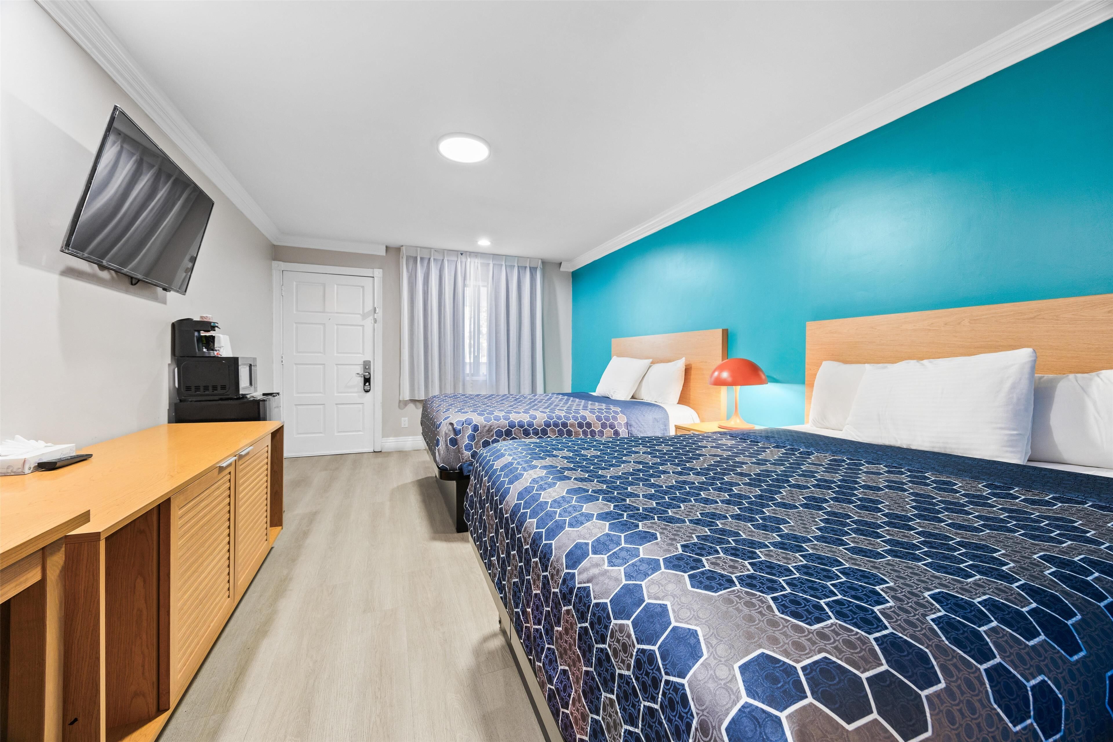 A hotel room with two beds, a desk with a coffee machine, a TV mounted on the wall, and a window with curtains.