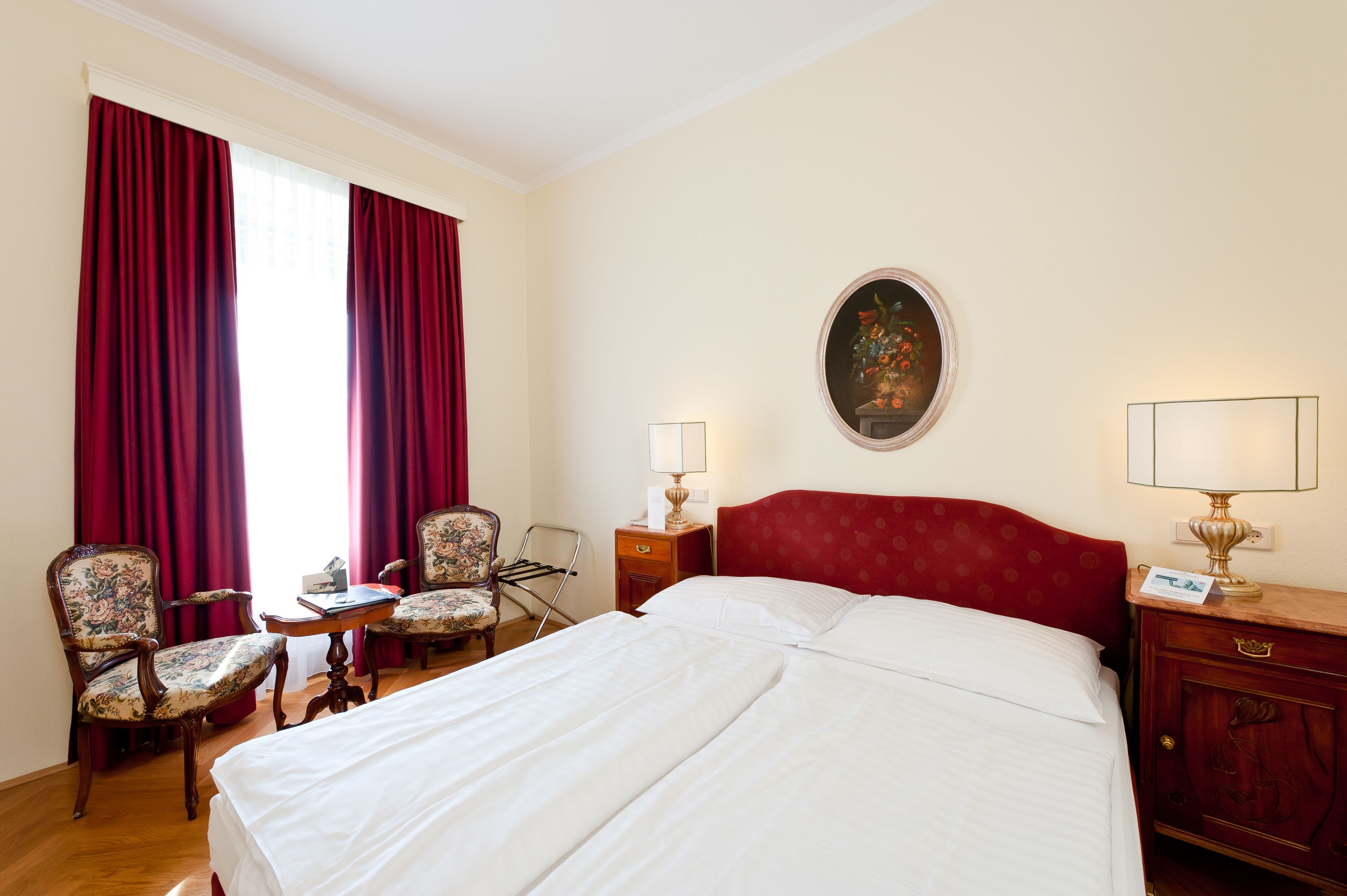 A hotel room with a large bed, two chairs, a small table, a nightstand with a lamp, and a painting on the wall.