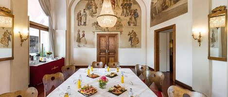 A dining room with a long table set for a meal, wooden chairs, a chandelier, and a mural on the wall.
