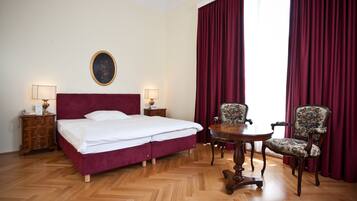 A hotel room with a large bed, two bedside tables with lamps, a round wooden table with chairs, and a large window with red curtains.