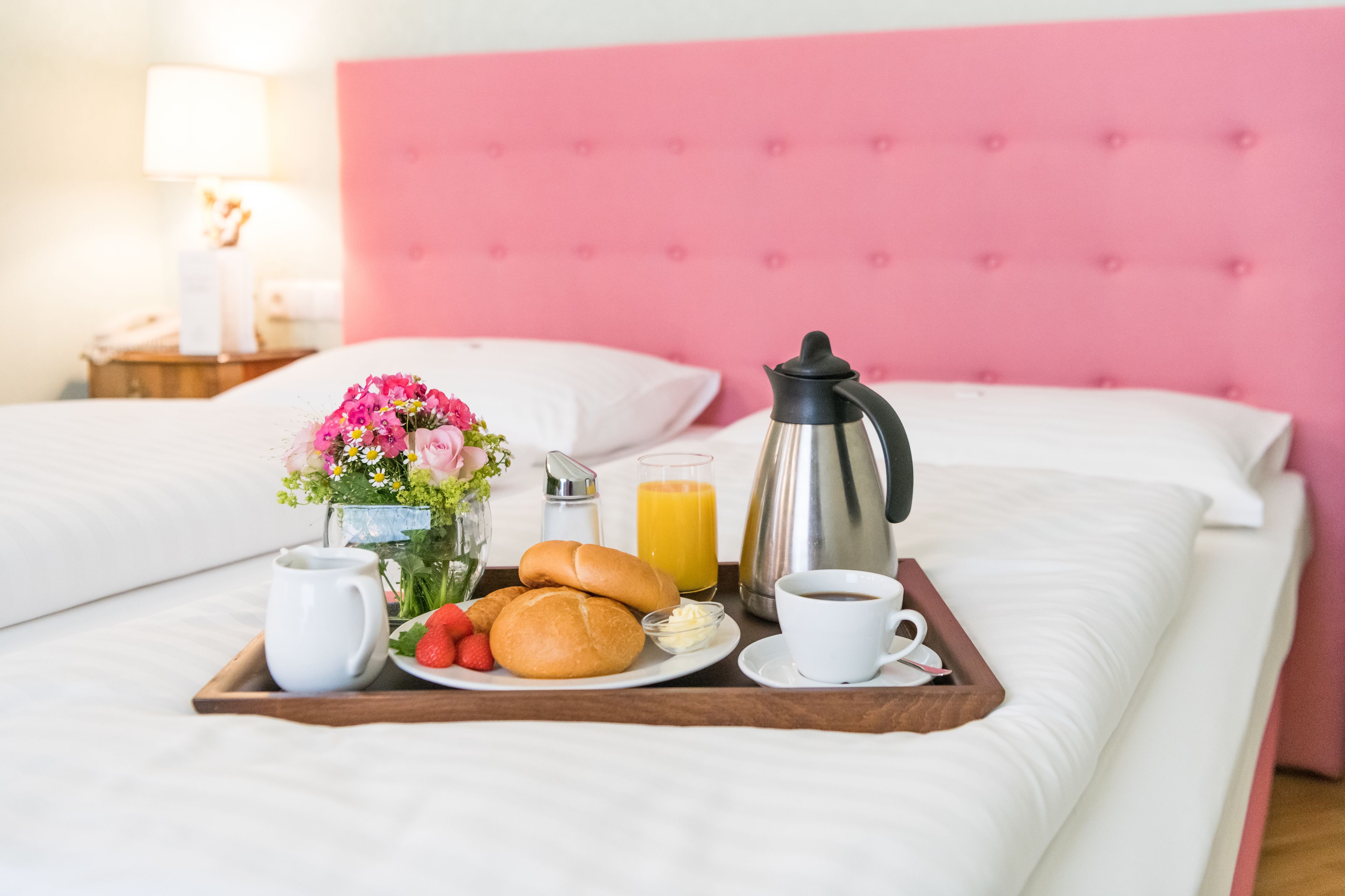 A neatly made breakfast tray on a bed with a pink headboard, including croissants, strawberries, a coffee cup, and a juice glass.