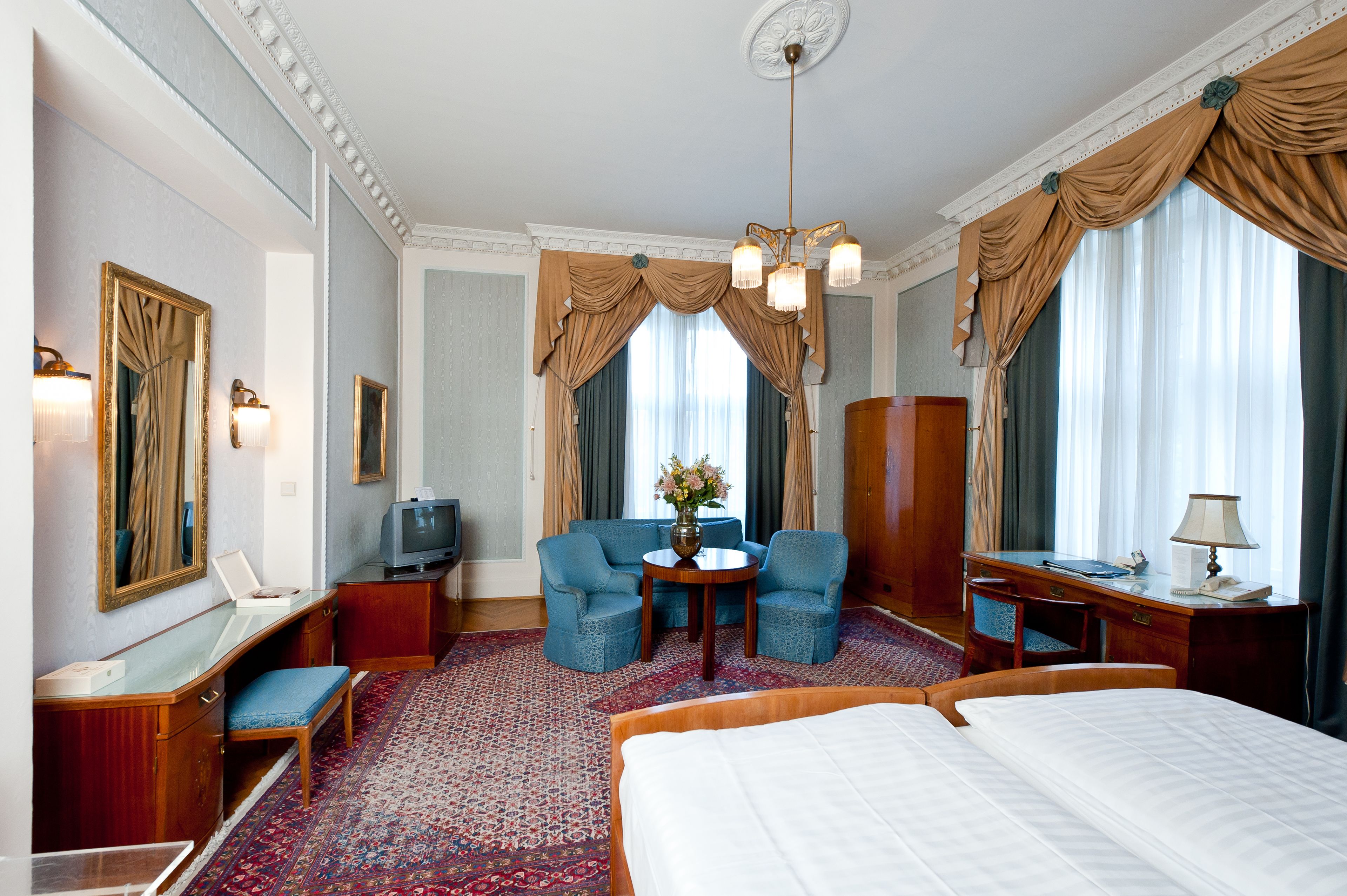 A hotel room with a large bed, two armchairs, a small table with a vase, a desk with a lamp, a television, and a large window with curtains.