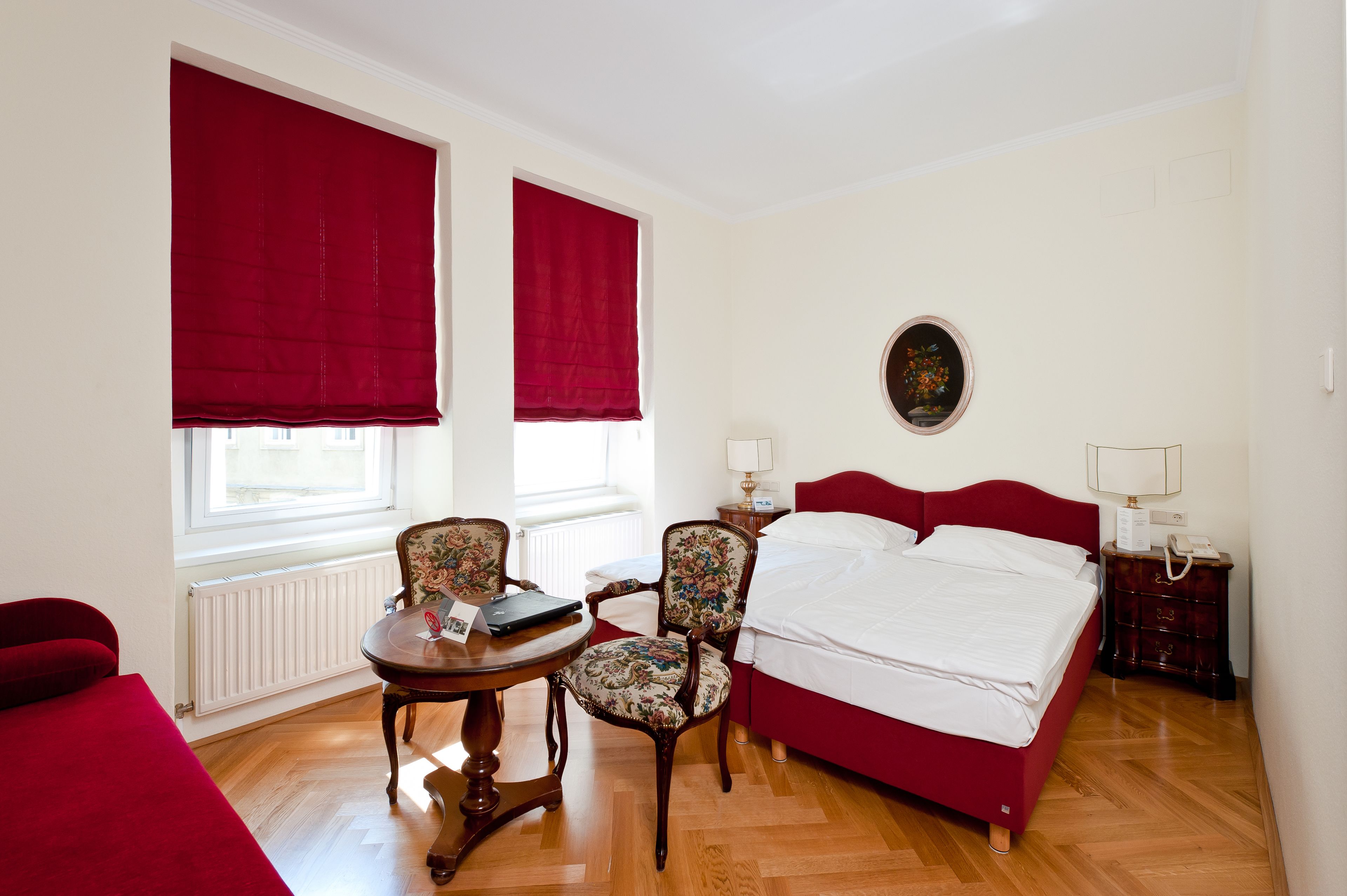 A hotel room with a bed, two chairs, a table, a lamp, and a window with red curtains.